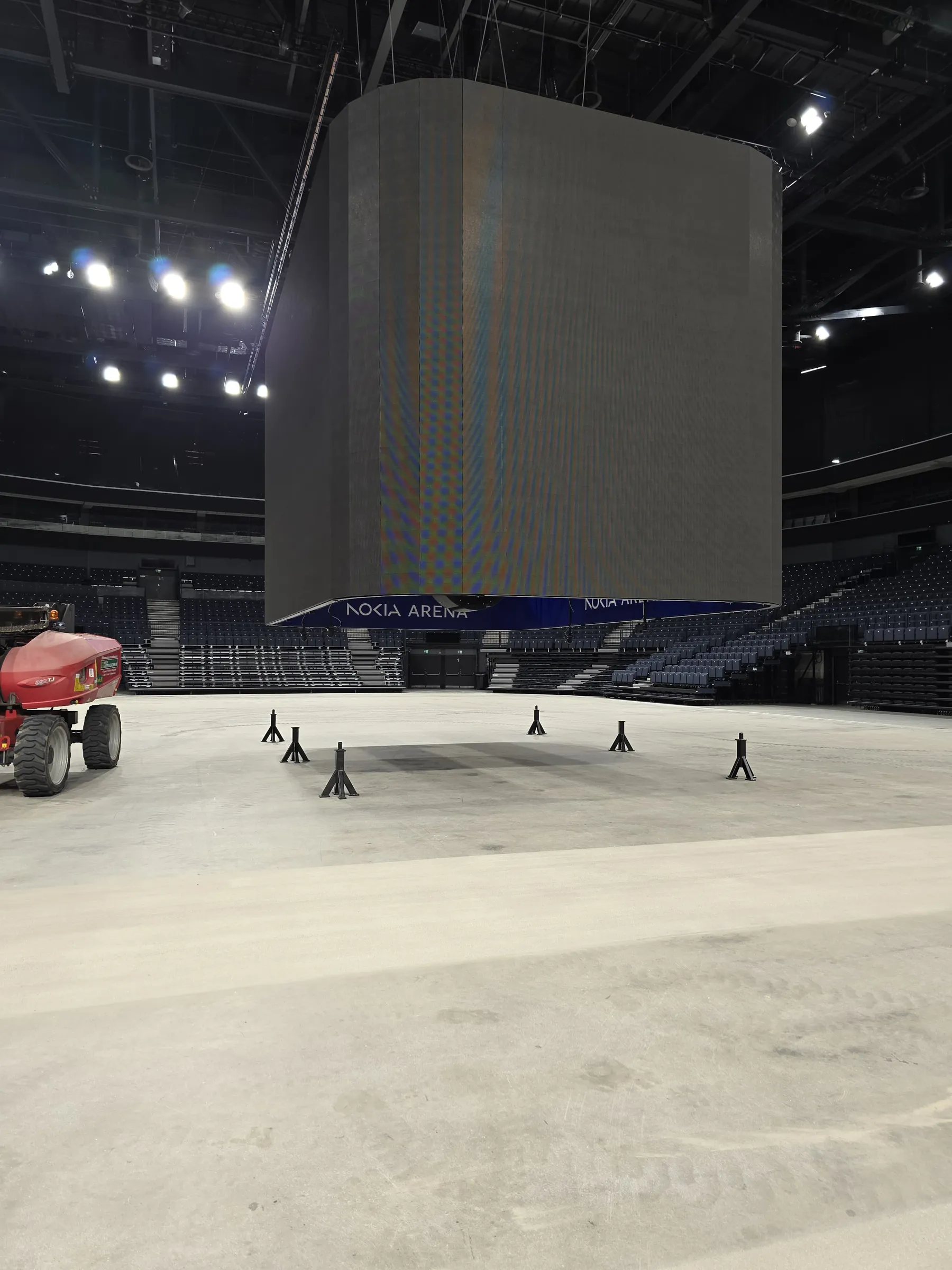 Suuri, tumma LED-näyttö roikkuu areenan keskellä. Sen alapuolella on sininen banneri tekstillä "NOKIA ARENA". Taustalla näkyy tummansinisiä istuinkatsomoita. Vaalealla betonilattialla on rengasjälkiä ja useita mustia, Y-muotoisia telineitä. Kuvan vasemmassa reunassa on osittain punainen työkone. Katon kirkkaat valot valaisevat areenaa.