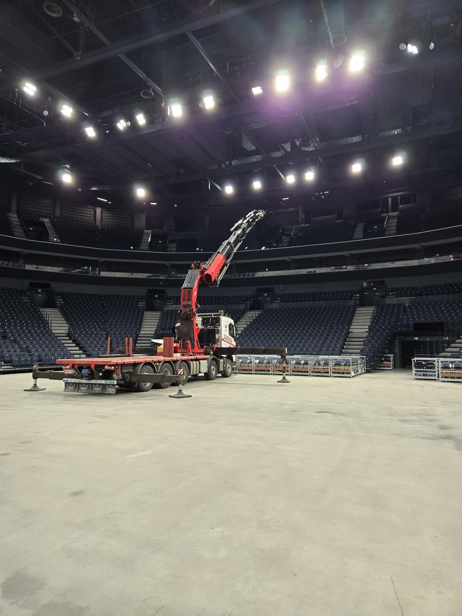 Suuri areena tai halli, jossa on tyhjiä tummansinisiä istuinrivejä. Vaalealla betonilattialla seisoo punavalkoinen kuorma-auto, jonka nosturivarsi on ojennettuna ylöspäin. Kuorma-auton tukijalat ovat levitettyinä. Taustalla istuinrivien edessä on pinottuna metallihäkkejä. Katon kirkkaat valot valaisevat tilaa.