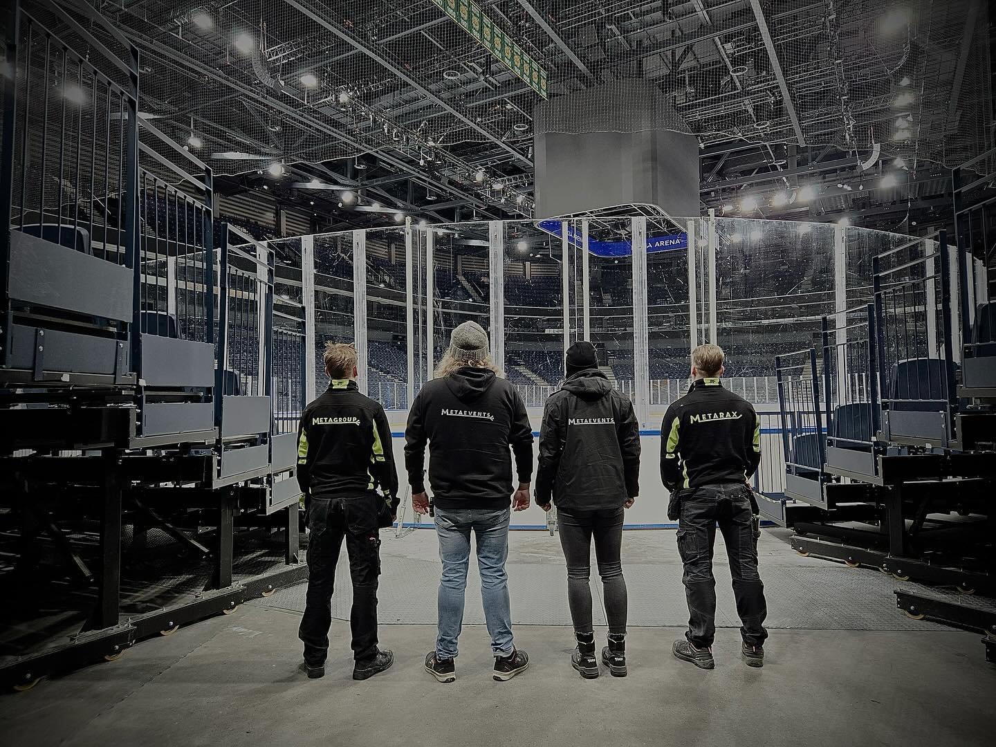 Neljä mustiin työtakkeihin pukeutunutta henkilöä seisoo selin kameraan avarassa, hämärästi valaistussa areenassa. Takkeihin on painettu logot 'METAGROUP', 'METAEVENTS' ja 'METARAX'. Taustalla näkyy tyhjiä istuinrivejä, läpinäkyviä lasiaitoja ja suuri tulostaulu, jossa lukee 'A ARENA'. Lattialla on pinottuja mustia metallirakenteita.