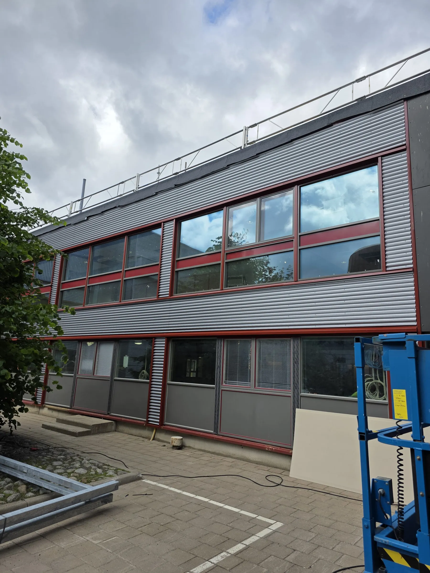 Kuva esittää kaksikerroksista rakennusta pilvisenä päivänä. Rakennuksen julkisivussa on harmaa aaltopeltiverhous ja punaiset ikkunankehykset. Ikkunoissa heijastuu pilvinen taivas. Rakennuksen edustalla on kiveystä, portaita, sininen työnostin oikealla ja maassa metallipalkkeja vasemmalla. Vasemmalla näkyy myös vihreitä puunlehtiä. Katolla on metallinen kaide.