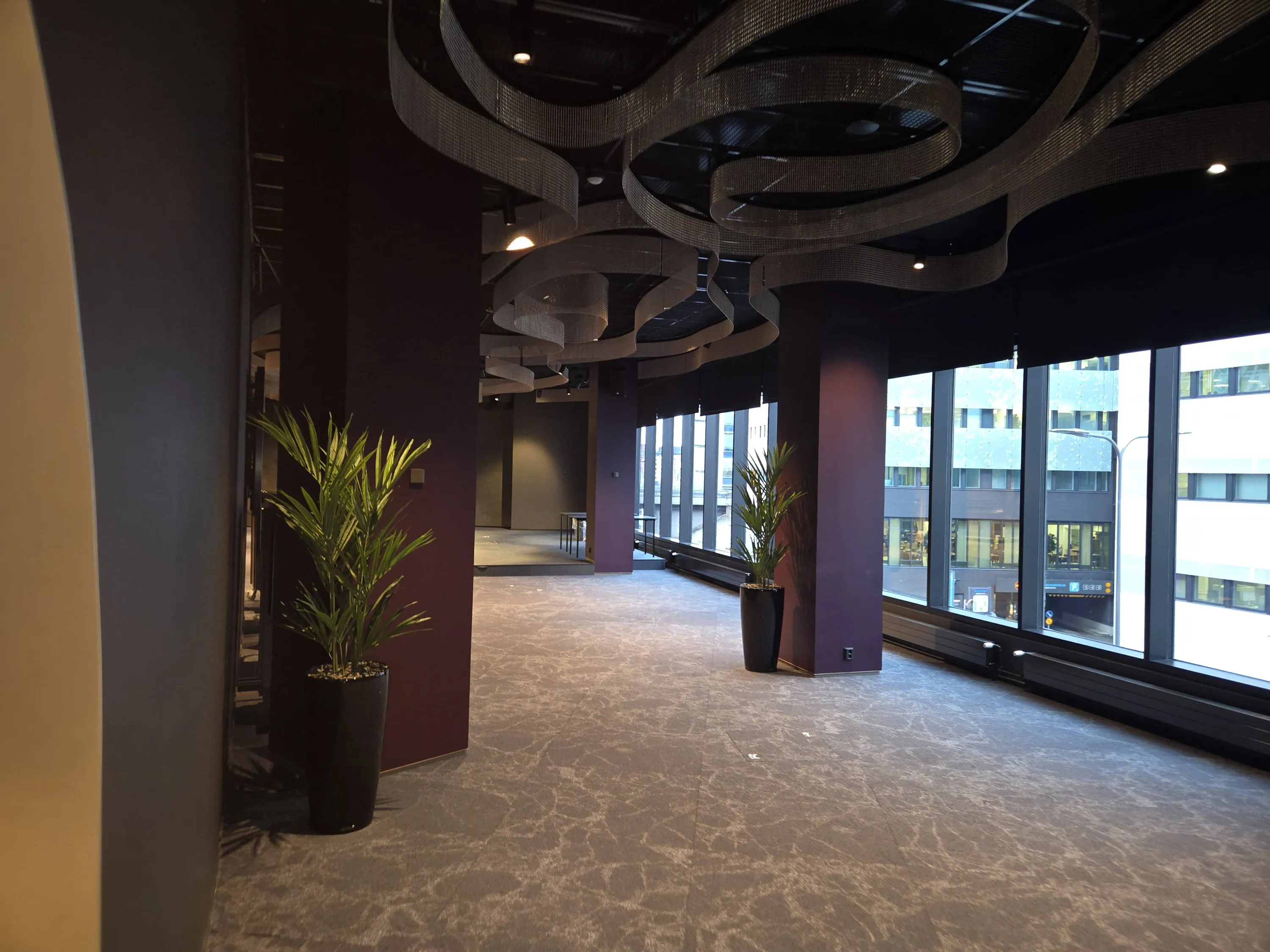 Tilava, moderni huone, jossa on tummanvioletteja pilareita ja vaaleanharmaa kuvioitu matto. Katossa on aaltoilevia metalliverkkorakenteita ja upotettuja spottivaloja. Huoneen oikealla puolella on suuret ikkunat, joista avautuu näkymä kaupungin rakennuksiin ja kadulle. Kaksi viherkasvia tummissa ruukuissa on sijoitettu pilareiden viereen. Valaistus tulee luonnonvalosta ikkunoista ja katon valaisimista.