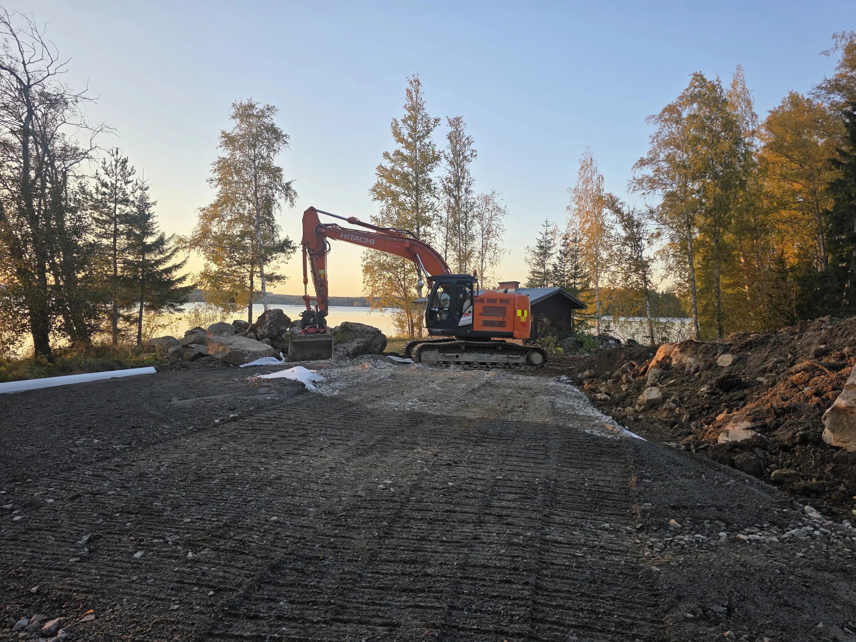 Oranssi Hitachi-kaivinkone seisoo tasoitetulla, tummalla maalla järven rannalla ilta-auringossa. Maassa näkyy telaketjun jälkiä ja vasemmalla on valkoinen putki. Kaivinkoneen edessä on kasa kiviä ja sen takana oikealla näkyy pieni puinen rakennus. Taustalla on tyyni järvi, jonka toisella rannalla on puita. Taivas on vaaleansininen ja puiden lehdet hehkuvat lämpimässä, kultaisessa valossa.