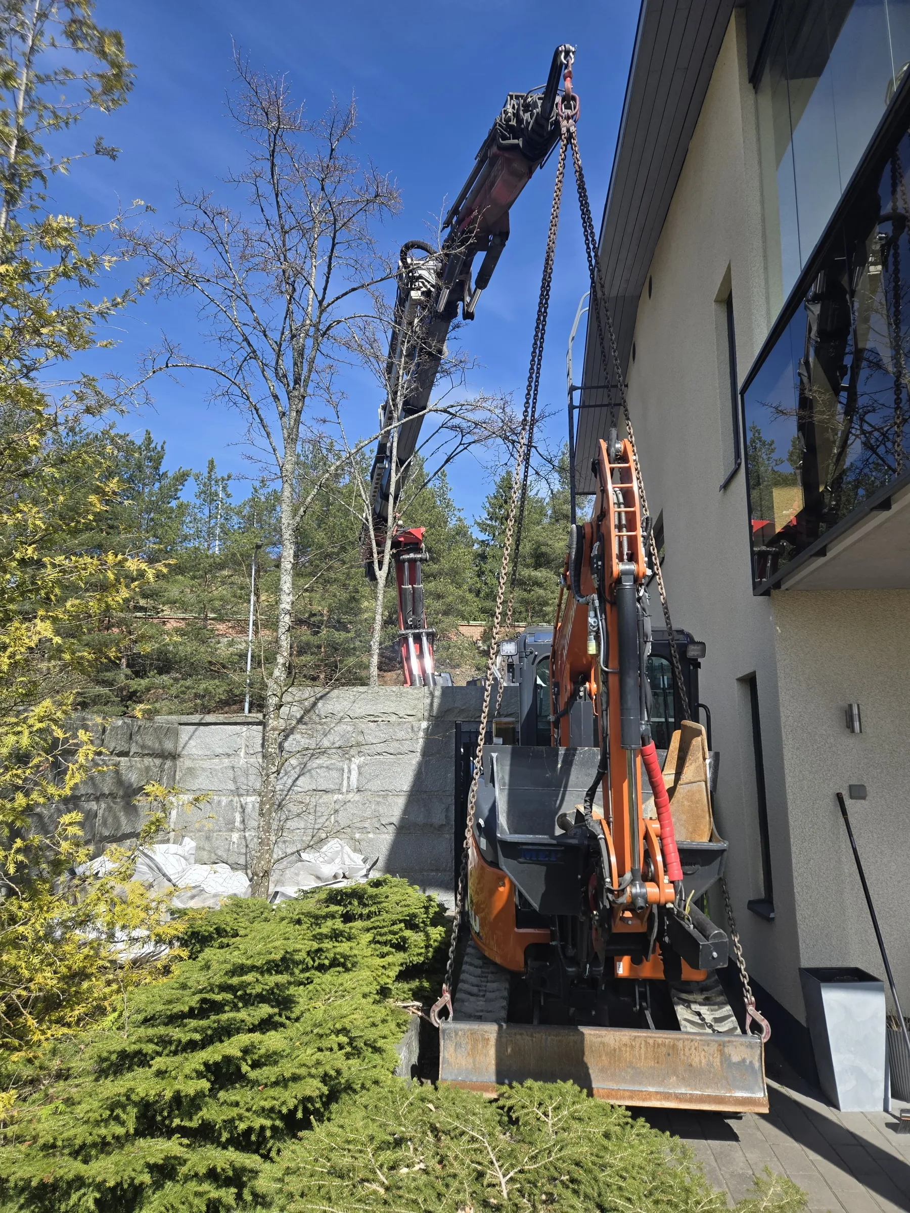Aurinkoisena päivänä oranssi kaivinkone nostetaan ketjuilla nosturin avulla vaalean rakennuksen viereen. Rakennuksessa on suuria ikkunoita, jotka heijastavat ympäristöä. Taustalla on lehdettömiä puita, havupuita ja harmaa kivimuuri. Kirkas sininen taivas näkyy ylhäällä.