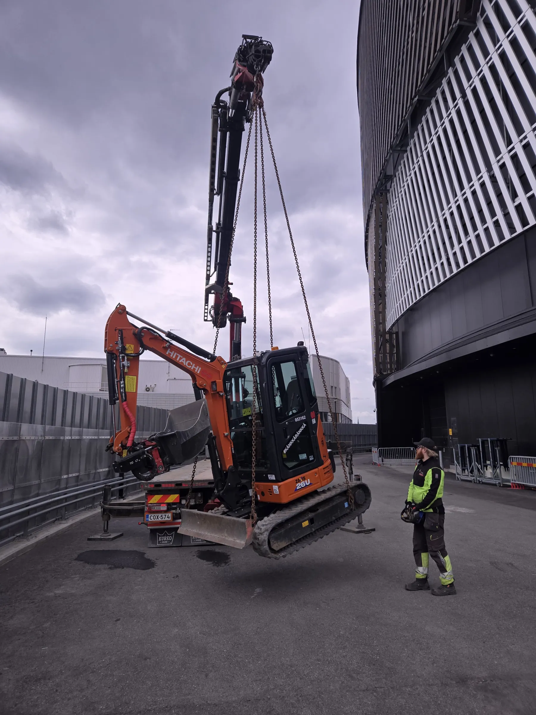 Oranssi Hitachi Zaxis 26U -minikaivinkone nostetaan kuorma-auton nosturilla pilvisenä päivänä. Kaivinkone roikkuu ilmassa ketjujen varassa. Oikealla puolella seisoo työntekijä limevihreässä ja mustassa työasussa, ohjaten nosturia kaukosäätimellä. Taustalla oikealla on moderni rakennus pystysuorilla valkoisilla säleillä ja vasemmalla harmaa seinä. Asfalttipinta on märkä paikoin.