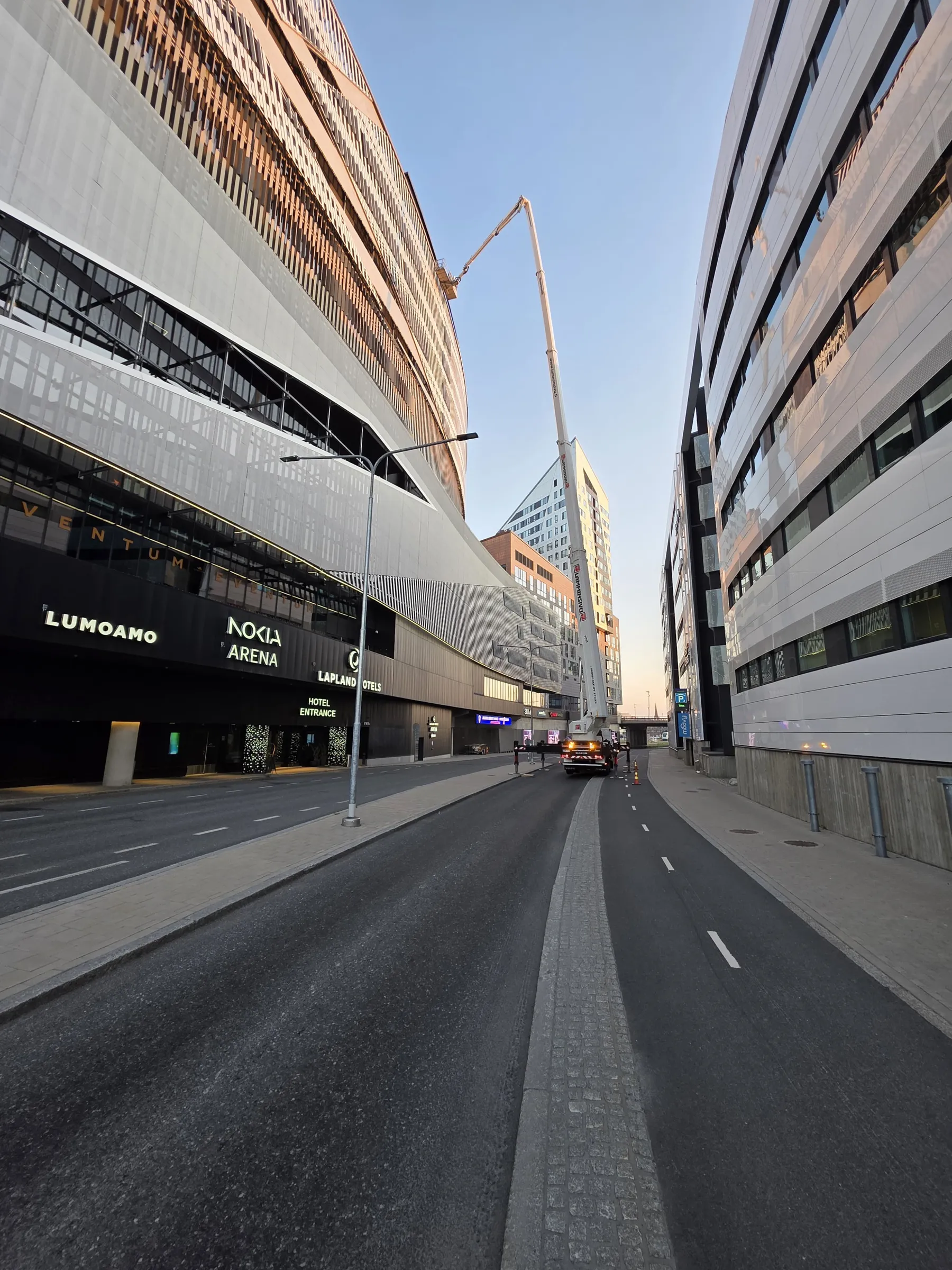 Katu kahden modernin rakennuksen välissä kirkkaassa päivänvalossa. Vasemmalla on kaareva rakennus, jonka alaosassa on musta julkisivu valaistuilla kylteillä 'LUMOAMO', 'NOKIA ARENA' ja 'LAPLAND HOTELS'. Yläosassa on ruskehtava, pystysuuntaisilla elementeillä varustettu julkisivu. Oikealla on valkoinen, ikkunariveillä varustettu rakennus. Keskellä katua on valkoinen nosturiauto, jonka puomi ulottuu vasemman rakennuksen yläosaan. Taivas on kirkas ja sininen.