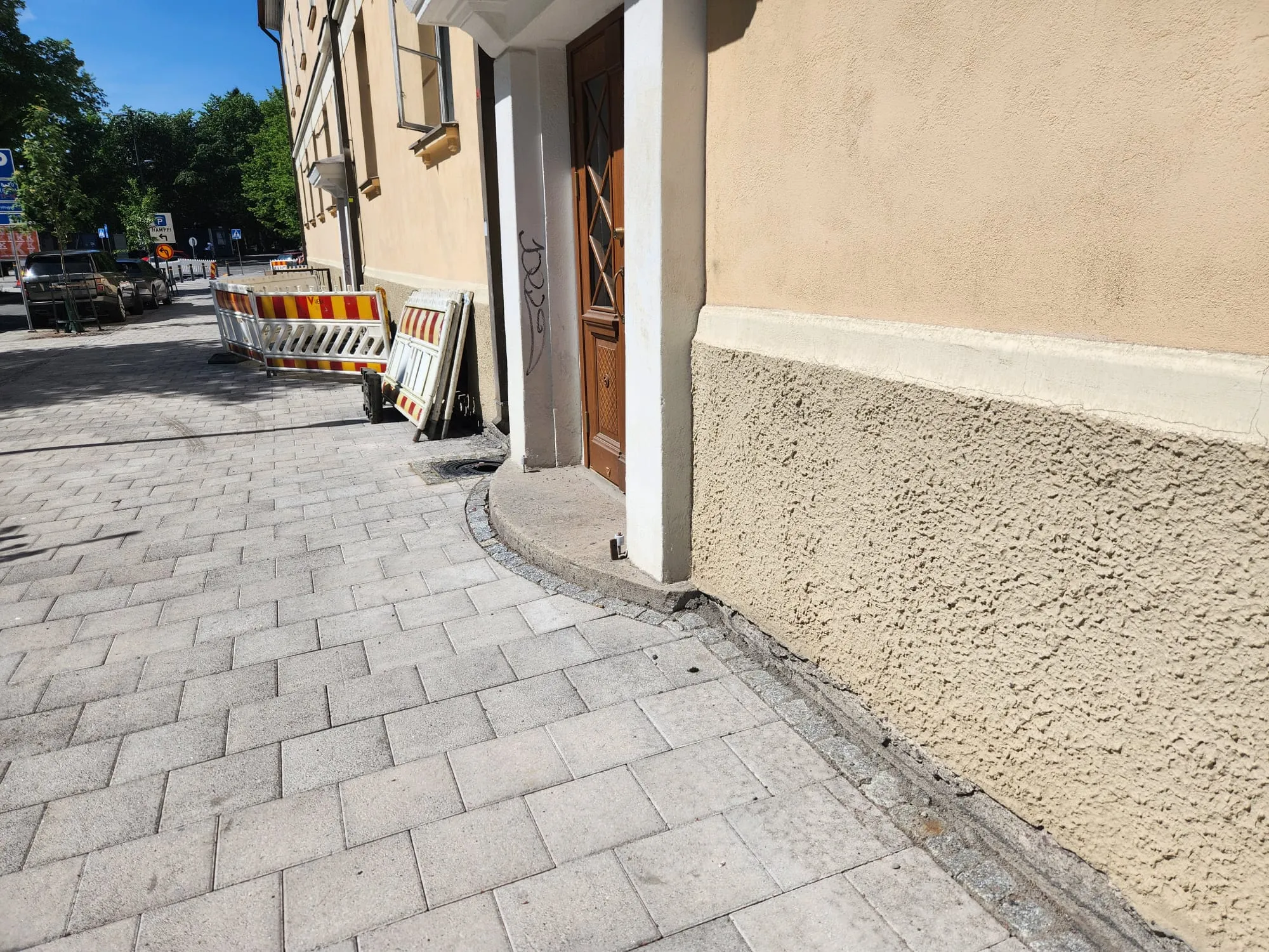 Aurinkoinen kuva jalkakäytävästä ja rakennuksen seinästä. Harmaat laattakivet peittävät jalkakäytävän. Oikealla on vaaleanbeigen värinen rakennuksen seinä, jonka alaosa on rosoista ja yläosa sileämpää. Ruskea puuovi, jota kehystävät valkoiset pylväät, on seinässä. Yhdessä pylväässä on mustia graffiteja. Kadulla näkyy punavalkoisia työmaa-aitoja, tummia autoja ja liikennemerkkejä. Taustalla on vihreitä puita ja sininen taivas.