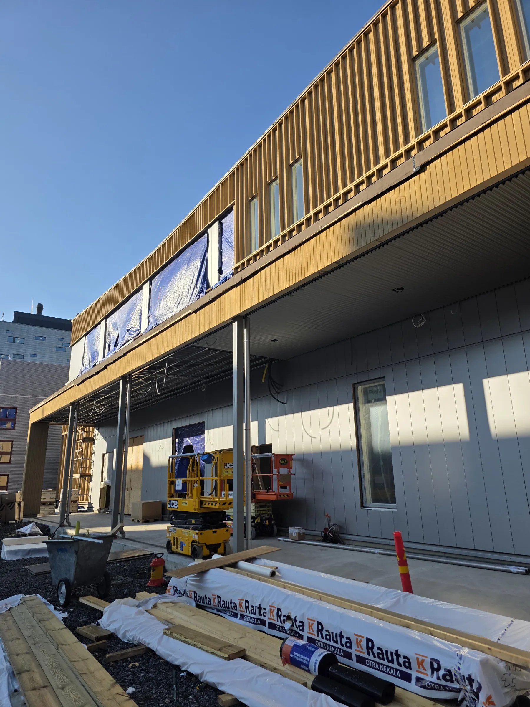 Rakennustyömaa aurinkoisena päivänä. Keskeneräinen rakennus, jonka yläosassa on pystysuorat vaaleanruskeat puurimat ja ikkunoita, joista osa on peitetty sinisillä suojapeitteillä. Rakennuksen alaosassa on harmaat paneelit ja ikkunoita. Edustalla on katettu käytävä hopeisella aaltopahvikatolla ja tukipylväillä. Maassa on puutavaraa, pakattuja rakennusmateriaaleja, joissa lukee "K Rauta OTRA NEKALA", sekä keltainen JCB- ja oranssi hrk.fi-saksinosturi. Taustalla näkyy toinen harmaa rakennus ja kirkas sininen taivas. Voimakkaat varjot lankeavat rakennuksen seinille.