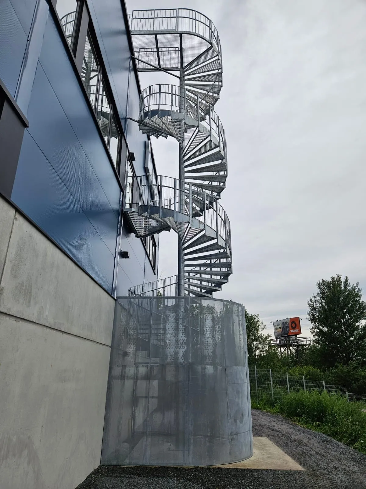 Modernin rakennuksen tummansininen, aaltopeltinen julkisivu ja harmaa betonijalusta. Rakennuksen sivulla on korkea, metallinen kierreportaikko verkkoportailla ja kaiteilla, joka alkaa sylinterimäisestä metallikotelosta. Taustalla näkyy pilvinen taivas, vihreitä puita ja kaukana oranssipohjainen mainostaulu.