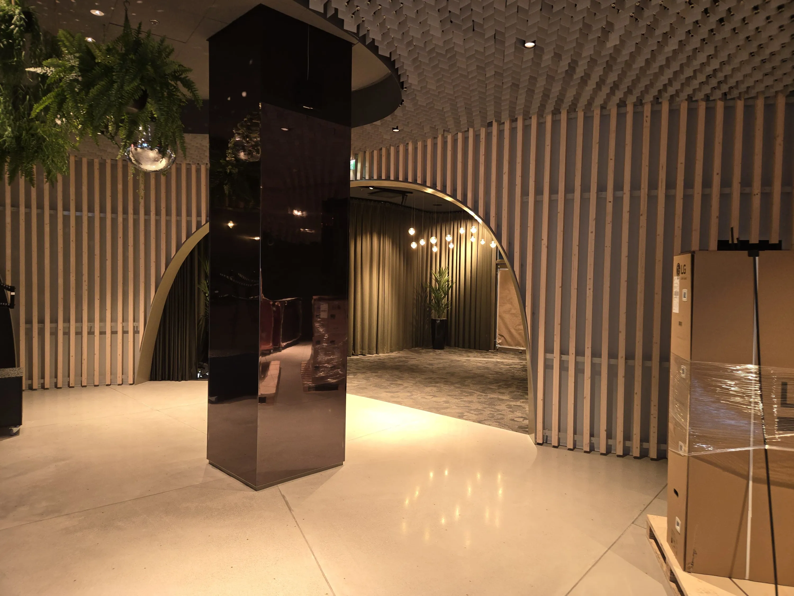 Moderni sisätila, jossa on tumma, heijastava pilari keskellä. Vasemmalla roikkuu vihreitä kasveja ja diskopallo. Seinillä on pystysuoria vaaleita puurimoja ja katossa on harmaa, chevron-kuvioitu laattapinta. Kaksi kaarevaa aukkoa johtaa taustalla olevaan tilaan, jossa näkyy verhoja, ruukkukasvi ja riippuvia pallovalaisimia. Oikealla on pahvilaatikoita lavalla, joissa on LG-logo. Lämmin valaistus luo tunnelmaa ja heijastuu kiiltäviltä pinnoilta.