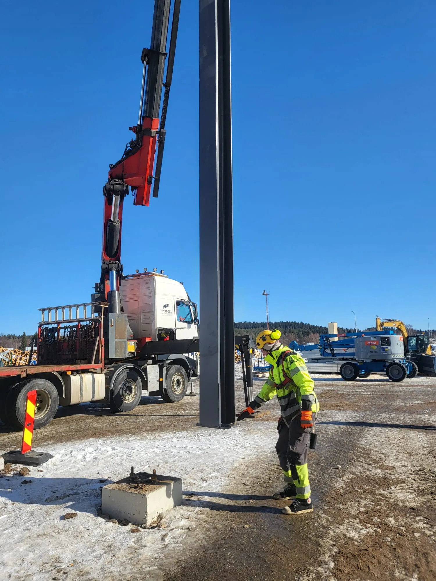 Työntekijä keltaisissa huomiovaatteissa ja kypärässä ohjaa tummaa metallipylvästä asennuspaikalle. Pylväs on punaisen nosturin varassa, joka kuuluu valkoiseen kuorma-autoon. Maassa on lunta ja jäätä. Taustalla näkyy puupinoja, sininen henkilönostin ja keltainen kaivinkone kirkkaan sinisen taivaan alla.