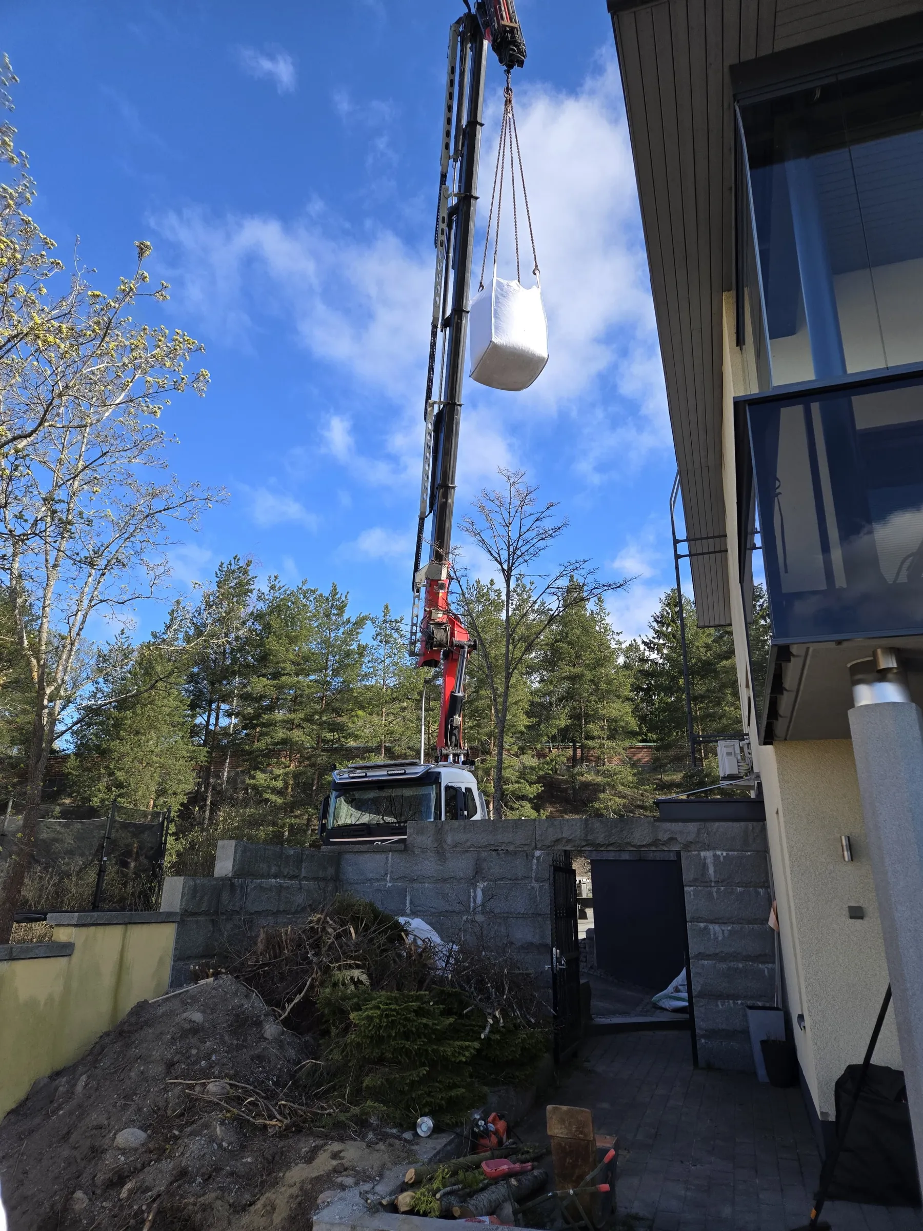 Nosturiauto nostaa valkoista suursäkkiä kirkkaan sinisellä, pilvisellä taivaalla. Kuvan vasemmalla puolella on lehtipuita ja taustalla mäntyjä. Oikealla näkyy modernin talon parveke ja suuria ikkunoita. Alhaalla on kivimuuri, multakasa ja katkaistuja puunrunkoja.