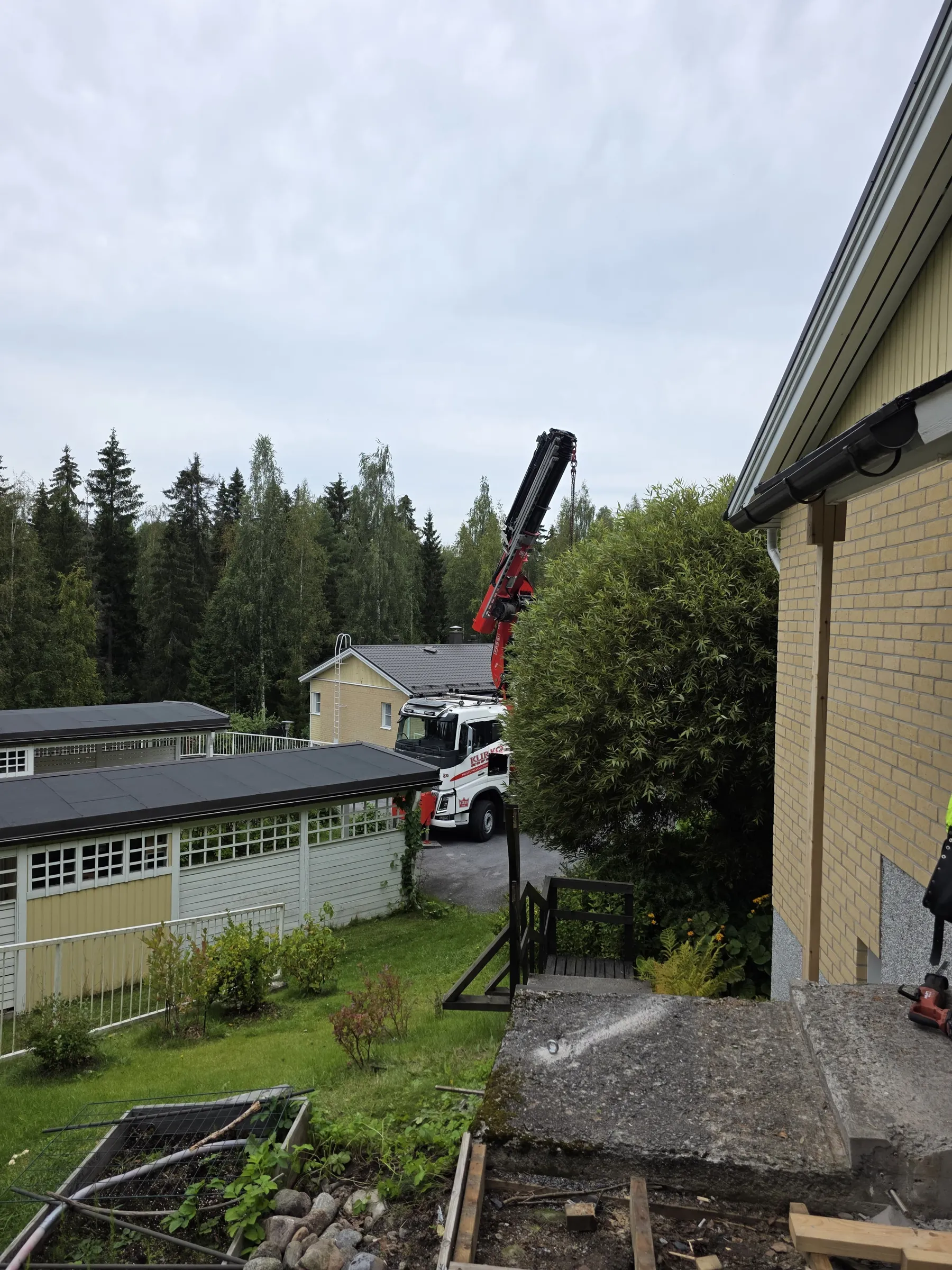 Pilvisenä päivänä otettu kuva rakennustyömaasta. Oikealla etualalla on keltainen tiilitalon seinä ja harmaa betonitasanne. Keskellä näkyy valkoinen nosturiauto, jonka punainen nosturivarsi on kohotettuna. Taustalla on tiheää metsää ja useita asuinrakennuksia, mukaan lukien keltainen talo tikkailla ja matalia rakennuksia tummilla katoilla ja valkoisilla ristikkoikkunoilla. Maassa on nurmikkoa ja puutarha-alue.