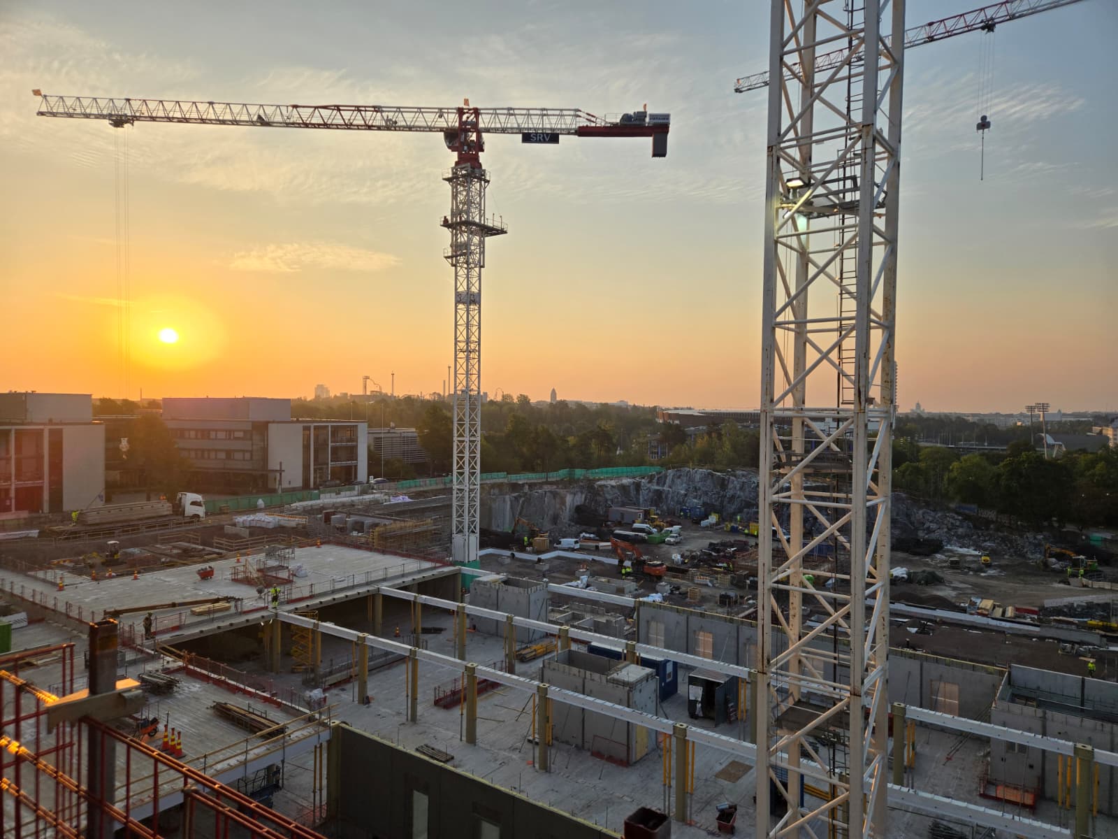 Rakennustyömaa auringonlaskun aikaan, jossa kaksi suurta torninosturia hallitsee näkymää. Aurinko laskee vasemmalla, värjäten taivaan oranssin ja keltaisen sävyihin. Etualalla on betonirakenteita, telineitä ja työntekijöitä. Taustalla näkyy kaupunkimaisema, vihreää kasvillisuutta ja kaivettu kallioalue.