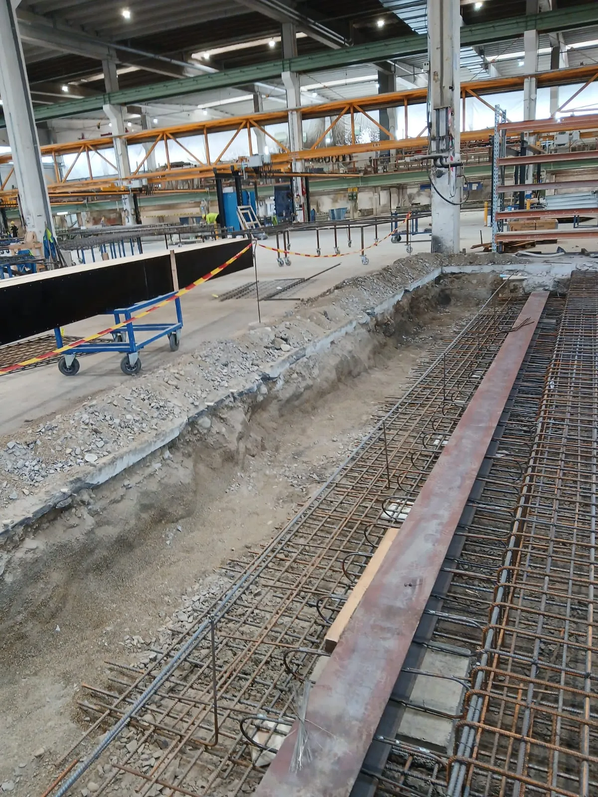 Kuva esittää sisätiloissa olevaa rakennustyömaata teollisuushallissa. Etualalla on kaivanto, jossa ja sen vieressä on laaja harjateräsraudoitus valmiina betonointia varten. Raudoituksen päällä on pitkä, ruosteinen metallipalkki. Keskialalla näkyy sininen kärry, jossa on musta paneeli ja kelta-punainen varoitusnauha. Taustalla on suuri halli, jossa on useita keltaisia siltanostureita ja työntekijöitä. Tila on kirkkaasti valaistu keinovalolla ja luonnonvalolla.