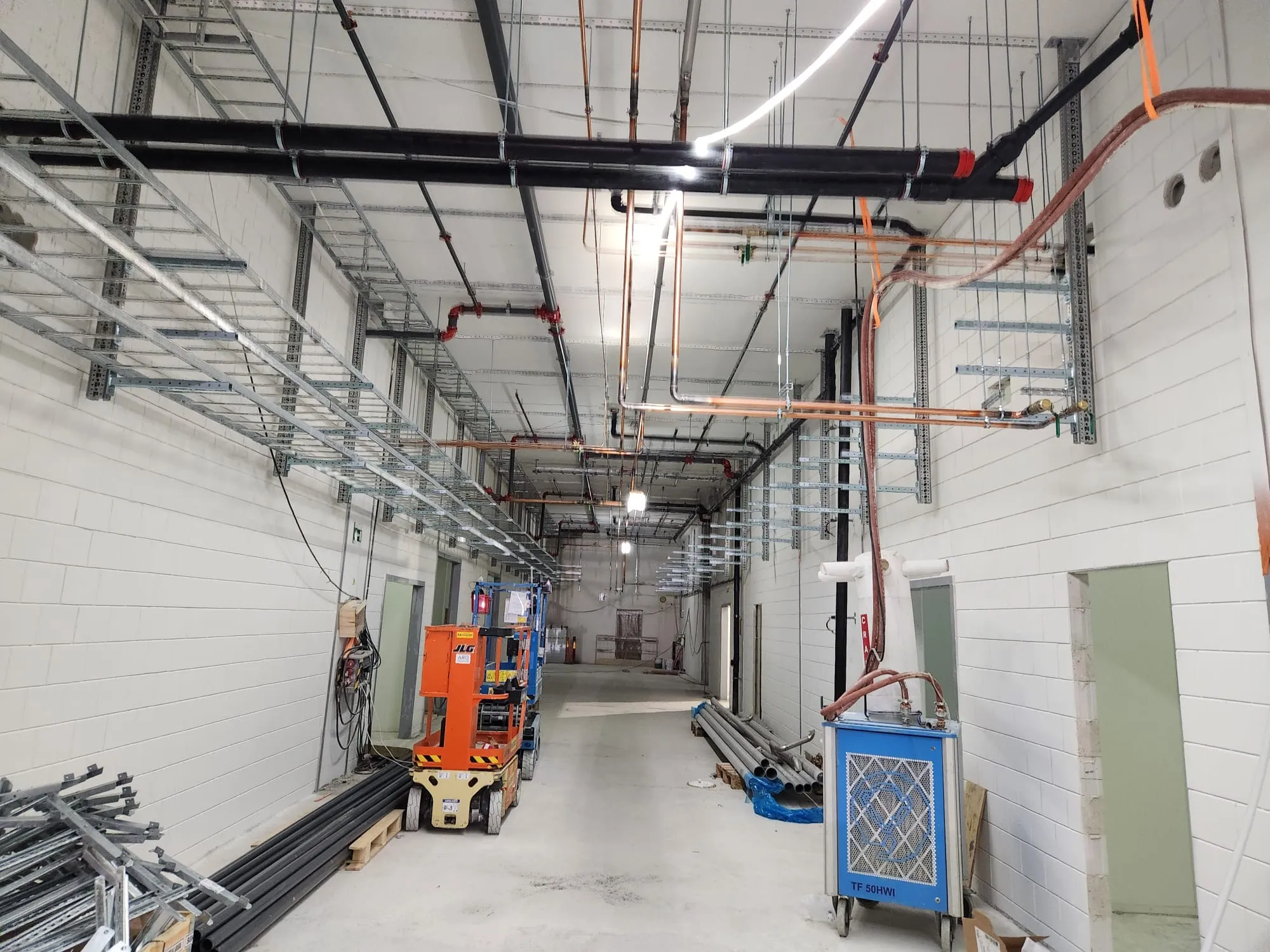 Pitkä, valkoisilla tiililohkoseinillä varustettu rakennustyömaan käytävä. Katossa kulkee runsaasti mustia ja kuparisia putkia sekä hopeisia kaapelihyllyjä. Lattialla on vaaleanharmaa betoni. Vasemmalla on kaksi oranssia JLG-henkilönostinta ja pinoja rakennusmateriaaleja, kuten metallitankoja ja mustia putkia. Oikealla on sininen pyörillä oleva kone ja pinoja harmaita putkia. Tila on kirkkaasti valaistu kattovaloilla.