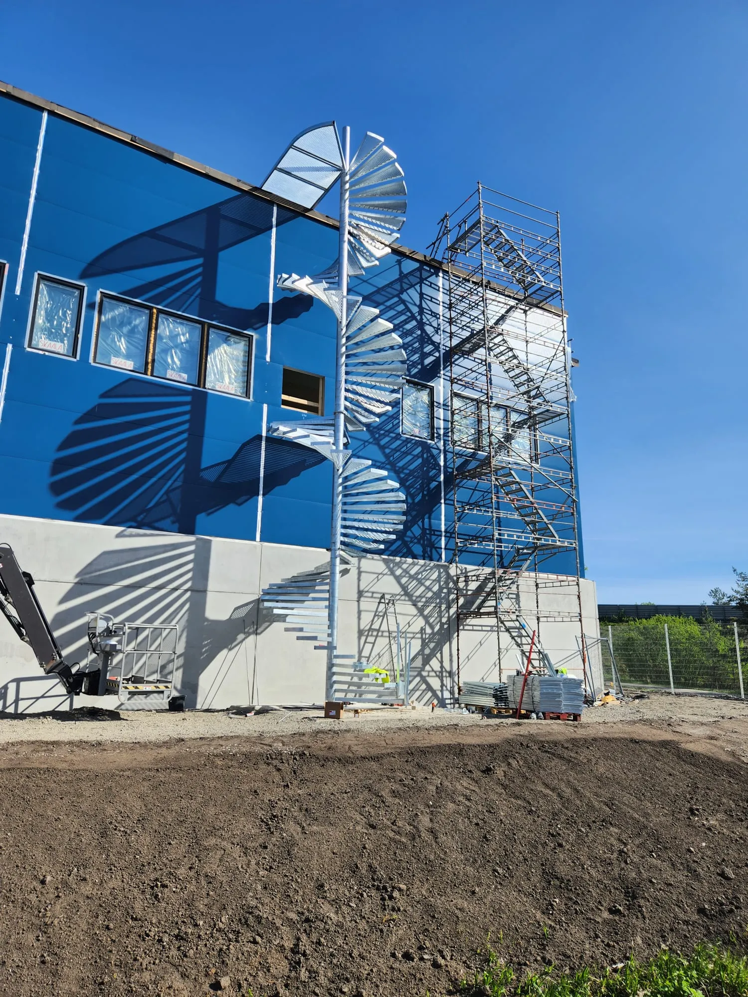 Rakenteilla oleva sininen rakennus kirkkaan sinisen taivaan alla. Rakennuksen vasemmalla puolella on hopeiset kierreportaat ja oikealla puolella rakennustelineet. Voimakas auringonvalo luo portaikon ja telineiden selkeät varjot siniselle seinälle ja harmaalle betonijalustalle. Etualalla on multakasa ja rakennustarvikkeita.