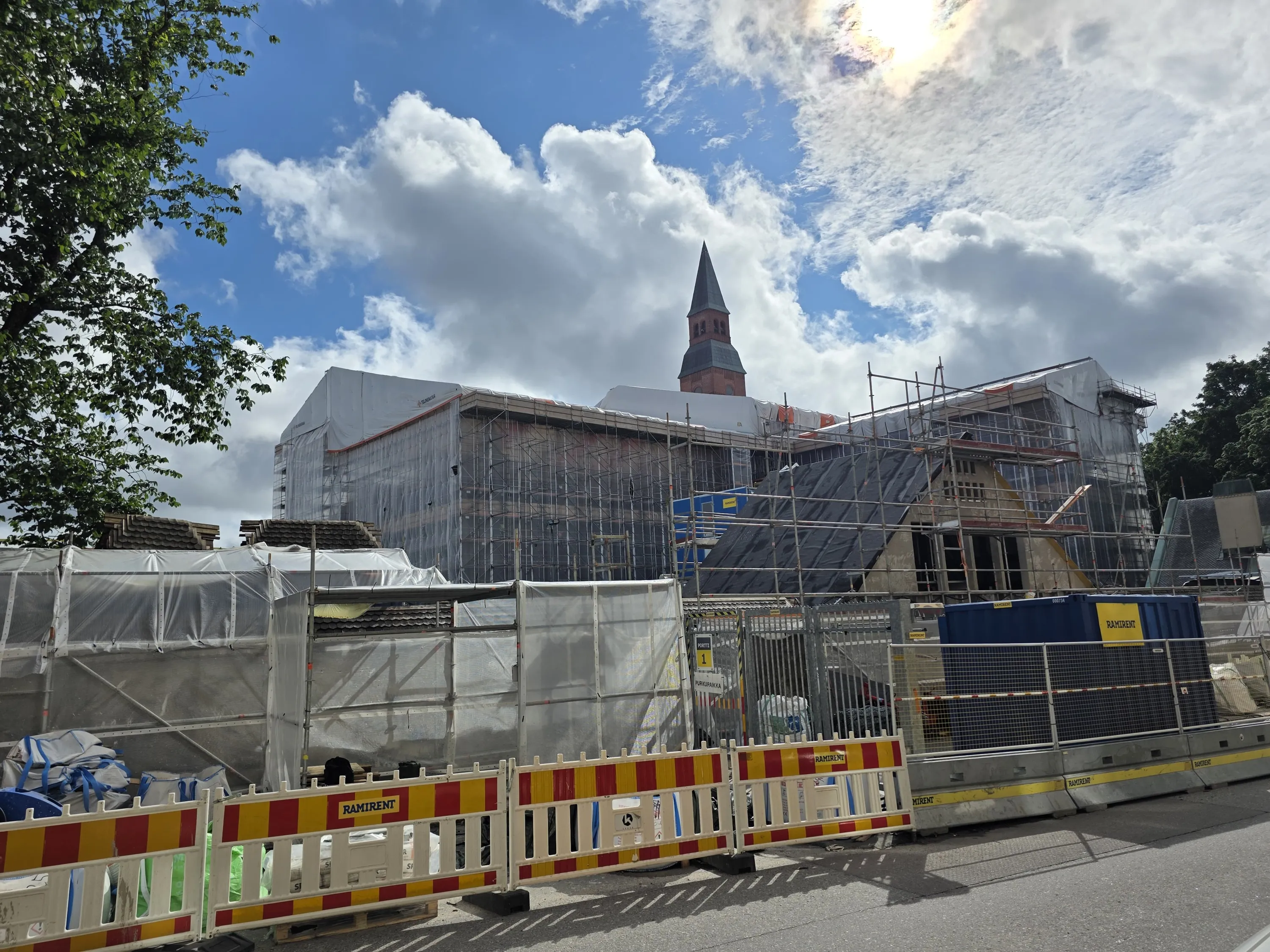Rakennustyömaa aurinkoisena päivänä. Suuri rakennus on peitetty telineillä ja valkoisilla suojapeitteillä. Taustalla kohoaa tiilinen kirkontorni tummalla huipulla. Vasemmalla näkyy vihreälehtinen puu. Etualalla on punakeltaisia ja harmaita rakennusaitoja. Taivas on sininen valkoisten ja harmaiden pilvien kanssa, ja aurinko paistaa kirkkaasti oikealta ylhäältä, luoden varjoja maahan.