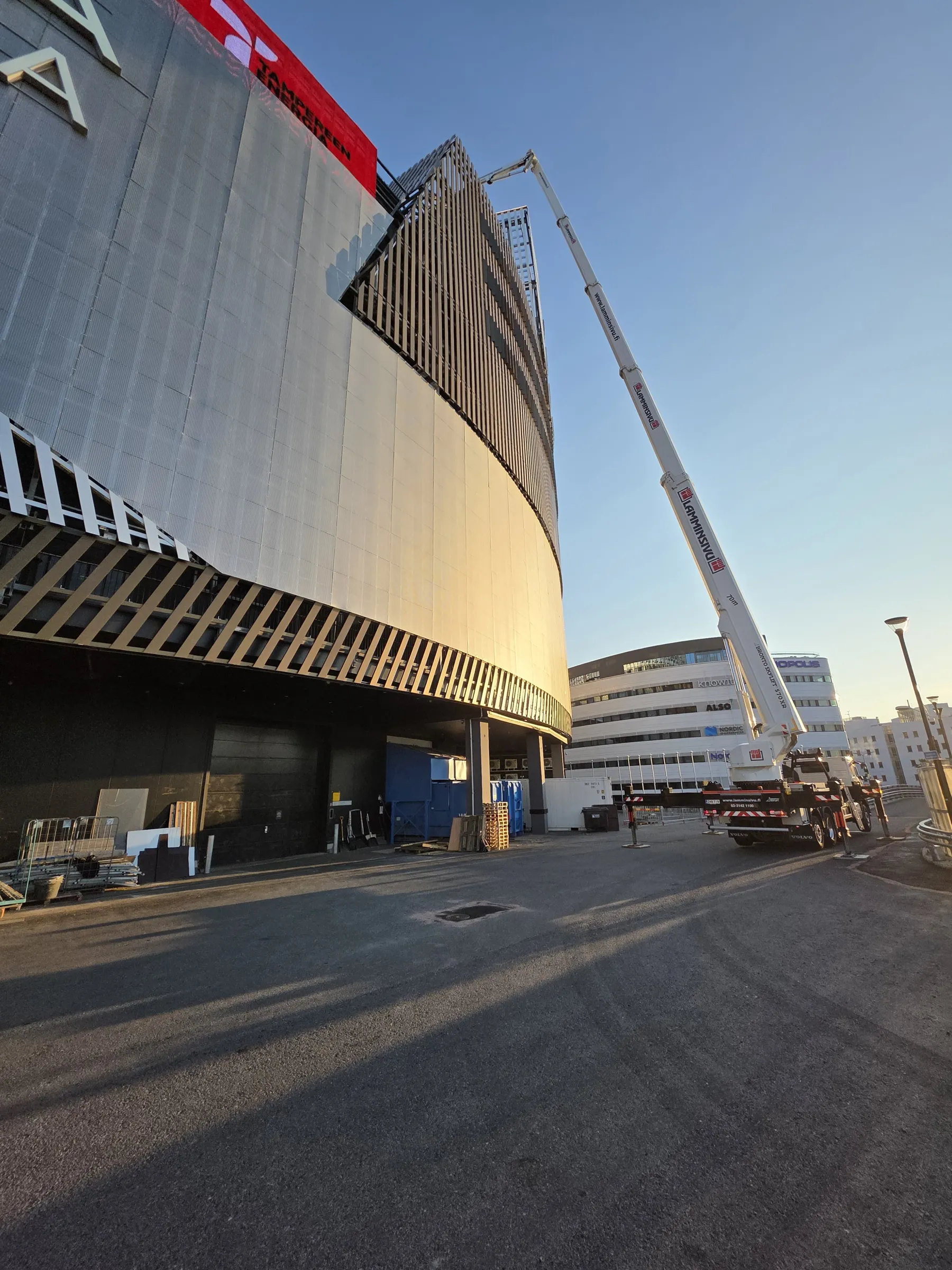 Suuri, kaareva rakennus, jossa on harmaita paneeleita ja puunvärisiä säleitä, nousee kuvan vasemmalla puolella. Rakennuksen yläosassa on punainen kyltti tekstillä "TAMPEREEN ENERGIA". Oikealla puolella valkoinen puominosturi ulottuu kohti rakennusta kirkkaan sinistä taivasta vasten. Maassa näkyy pitkiä varjoja ja rakennustarvikkeita. Taustalla on toinen valkoinen rakennus.