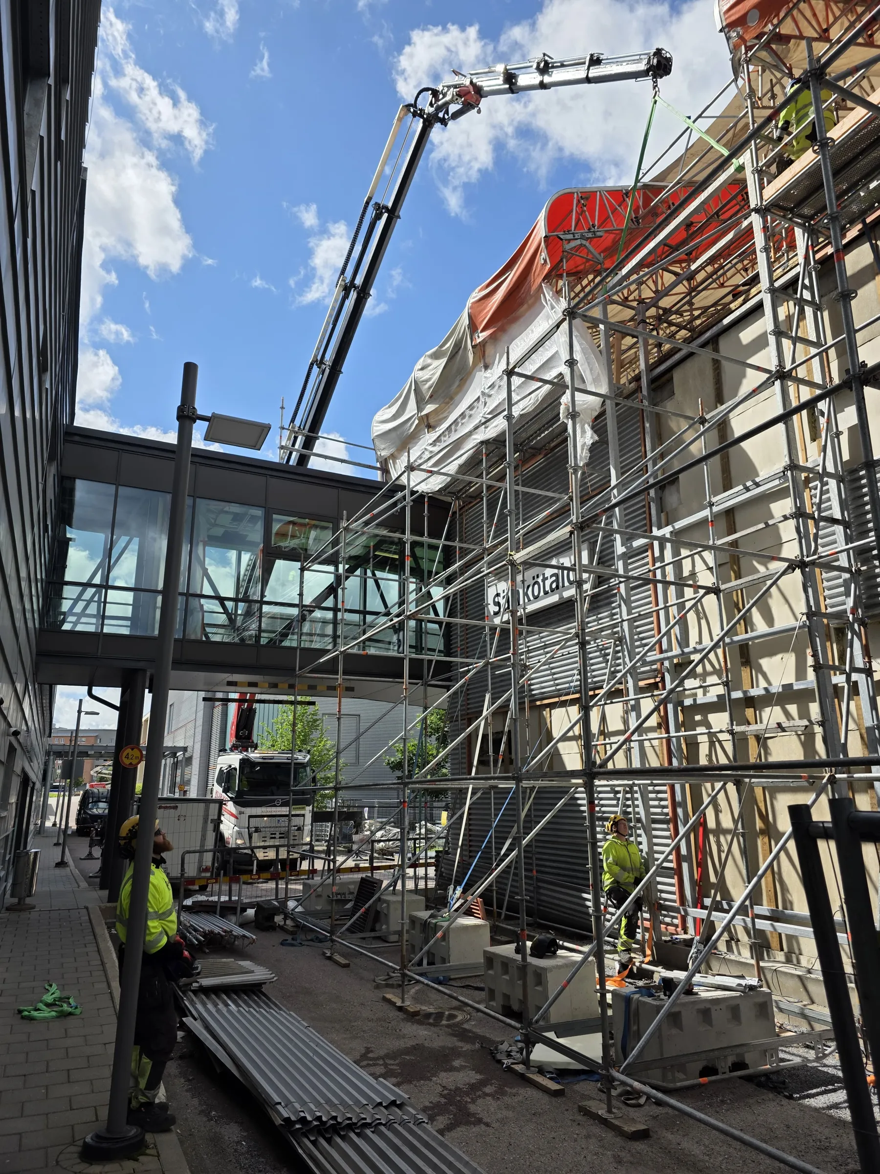 Aurinkoinen kuva rakennustyömaasta, jossa betonipumpun puomi ulottuu rakennustelineiden ja pressujen peittämän rakennuksen ylle. Taustalla sininen taivas valkoisine pilvineen. Vasemmalla on moderni rakennus lasi-ikkunoineen. Oikealla oleva rakennus on laajojen hopeanväristen telineiden ympäröimä, ja sen seinässä lukee "Sähkötalo". Maassa on rakennusmateriaaleja ja kaksi keltaisiin huomioliiveihin pukeutunutta työntekijää katsoo ylös.