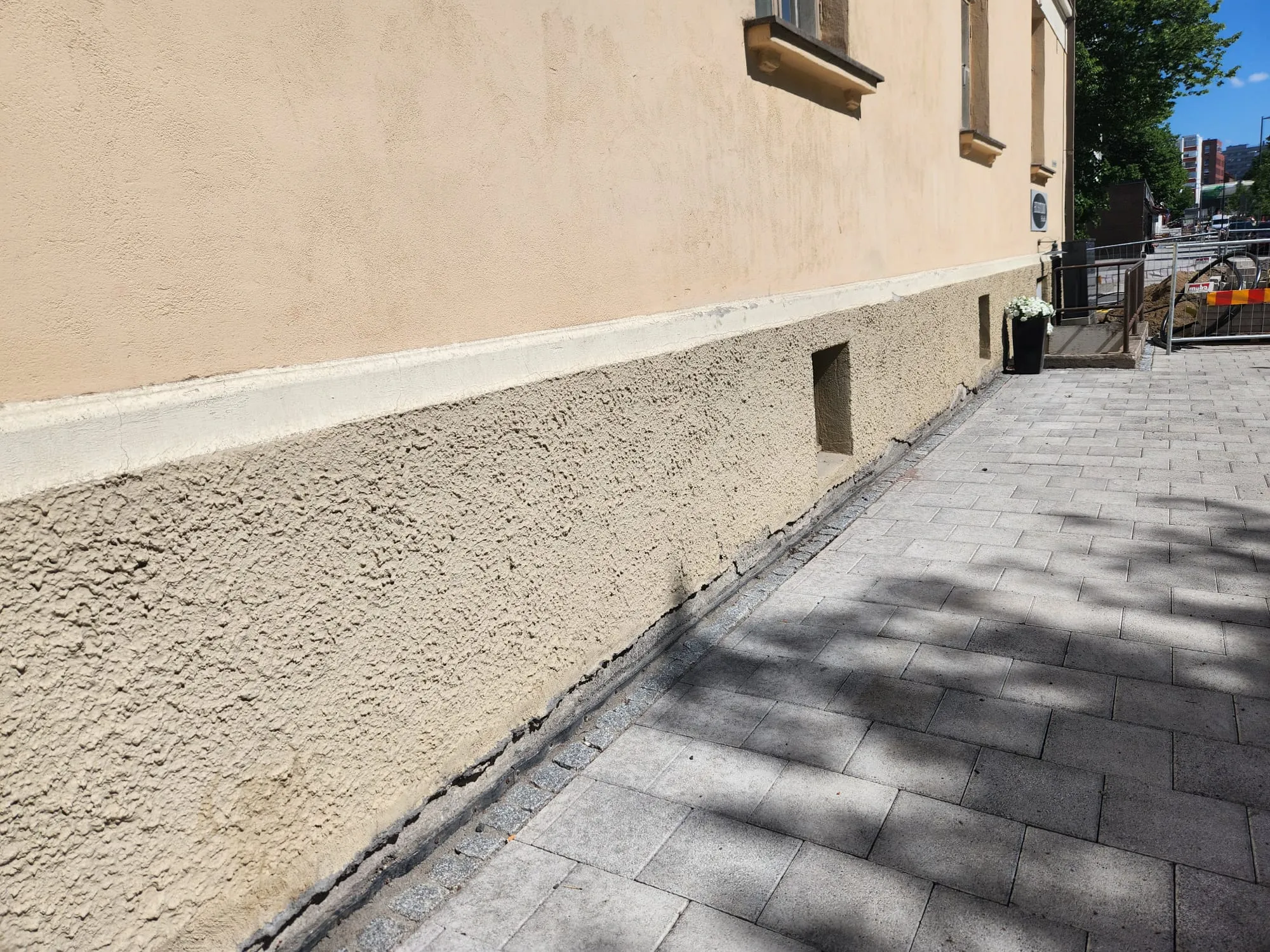 Aurinkoinen kuva rakennuksen vaaleanbeigestä seinästä ja harmaasta jalkakäytävästä. Seinän yläosa on sileää rappausta, alaosa karkeampaa, hieman tummempaa beigenväristä sokkelia. Niiden välissä on valkoinen lista. Sokkelissa on kaksi pientä suorakaiteen muotoista aukkoa. Oikealla on musta kukkaruukku valkoisine kukkineen. Jalkakäytävä on harmaista päällystekivistä, ja siinä näkyy varjoja. Seinän juurella on pieniä mukulakiviä ja tummia halkeamia. Taustalla näkyy rakennustyömaa ja puita.