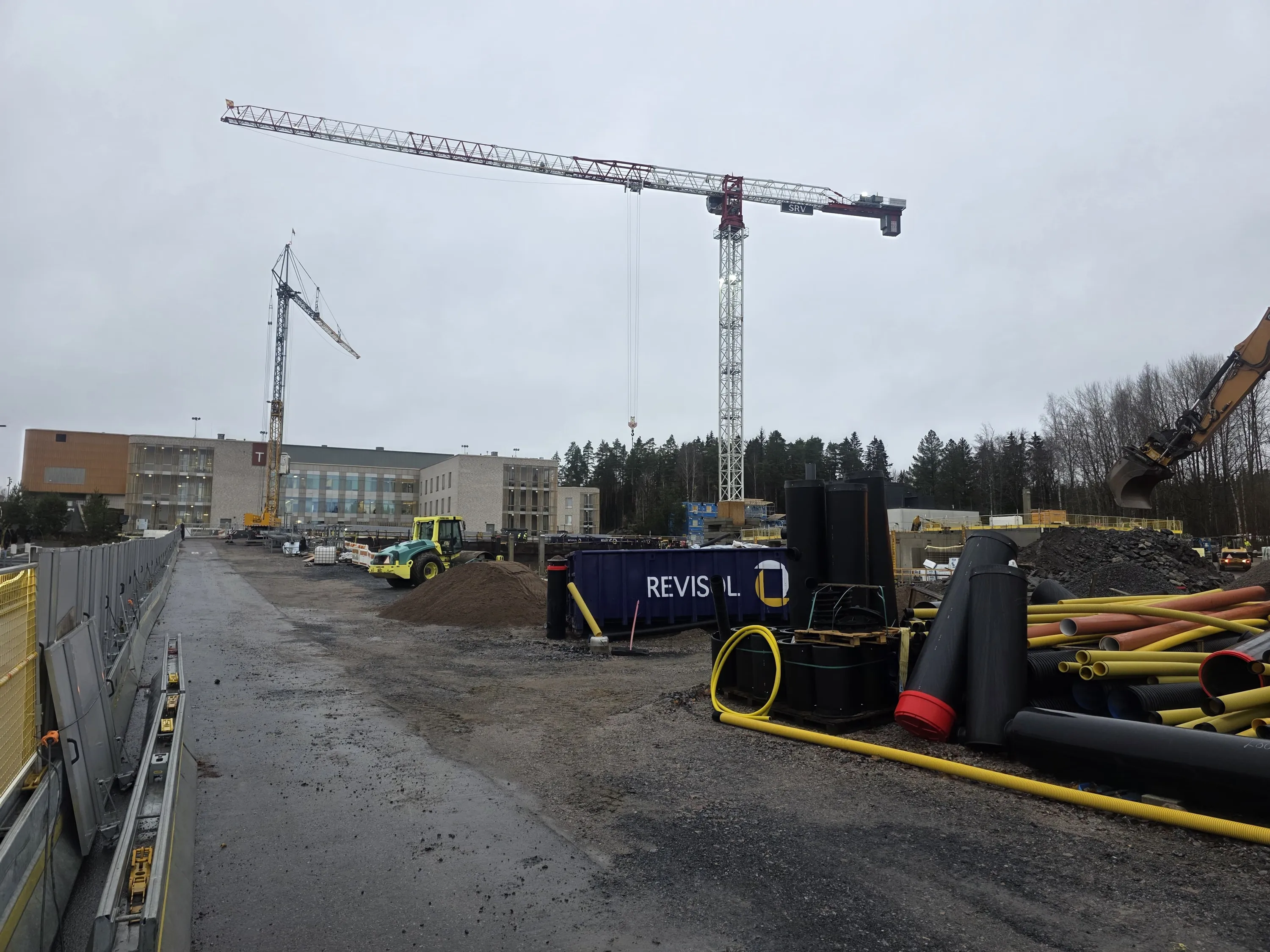Rakennustyömaa pilvisenä päivänä. Kaksi nostokurkea, joista etummaisessa lukee 'SRV', kohoaa taivaalle. Keskellä on rakenteilla oleva rakennus, jossa on puunvärisiä ja vaaleita kivipintoja. Edustalla on keltainen ja vihreä tiejyrä, hiekkakasa, sininen 'REVISOL'-kontti ja kasa erivärisiä putkia (keltaisia, mustia, oransseja). Oikealla näkyy kaivinkoneen kauha ja kivikasa. Maasto on märkää ja soraista, ja taustalla näkyy metsää. Valaistus on tasainen ja harmaa.