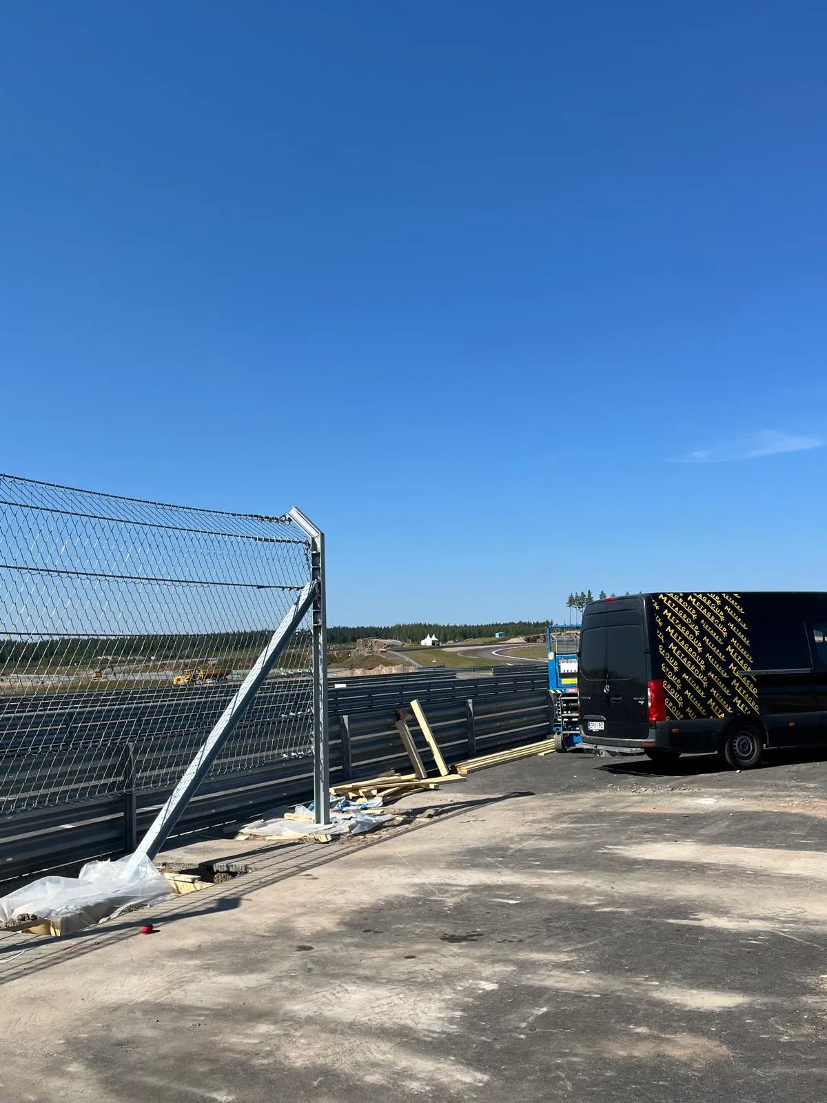 Kirkas sininen taivas hallitsee kuvaa. Etualalla vasemmalla on metallinen verkkoaita ja sen juurella rakennustarvikkeita. Oikealla on musta pakettiauto, jonka kyljessä on keltaisia "METAGROUP"-tekstejä. Taustalla mutkittelee kilparata metsäisessä maisemassa, jossa näkyy valkoisia telttoja. Maaperä on likaisen näköistä betonia ja maata aurinkoisessa säässä.