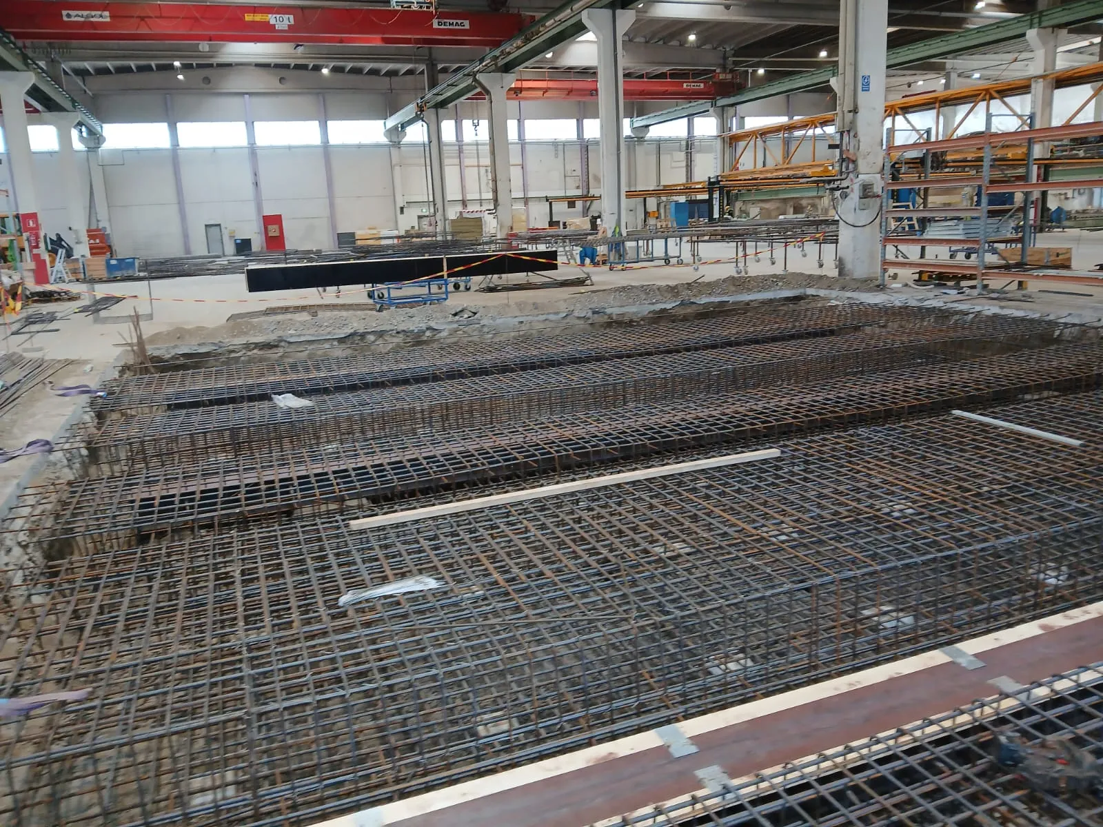 Sisätiloissa otettu kuva rakennustyömaasta. Etualalla on useita suuria, suorakaiteen muotoisia kaivantoja, jotka on täytetty ruosteisilla raudoitusverkoilla valmiina betonointiin. Taustalla näkyy laaja teollisuushalli, jossa on betonilattia, korkeat seinät ja yläpuolella punaisia siltanostureita sekä keltaisia tukirakenteita. Valaistus on kirkas ja tasainen.