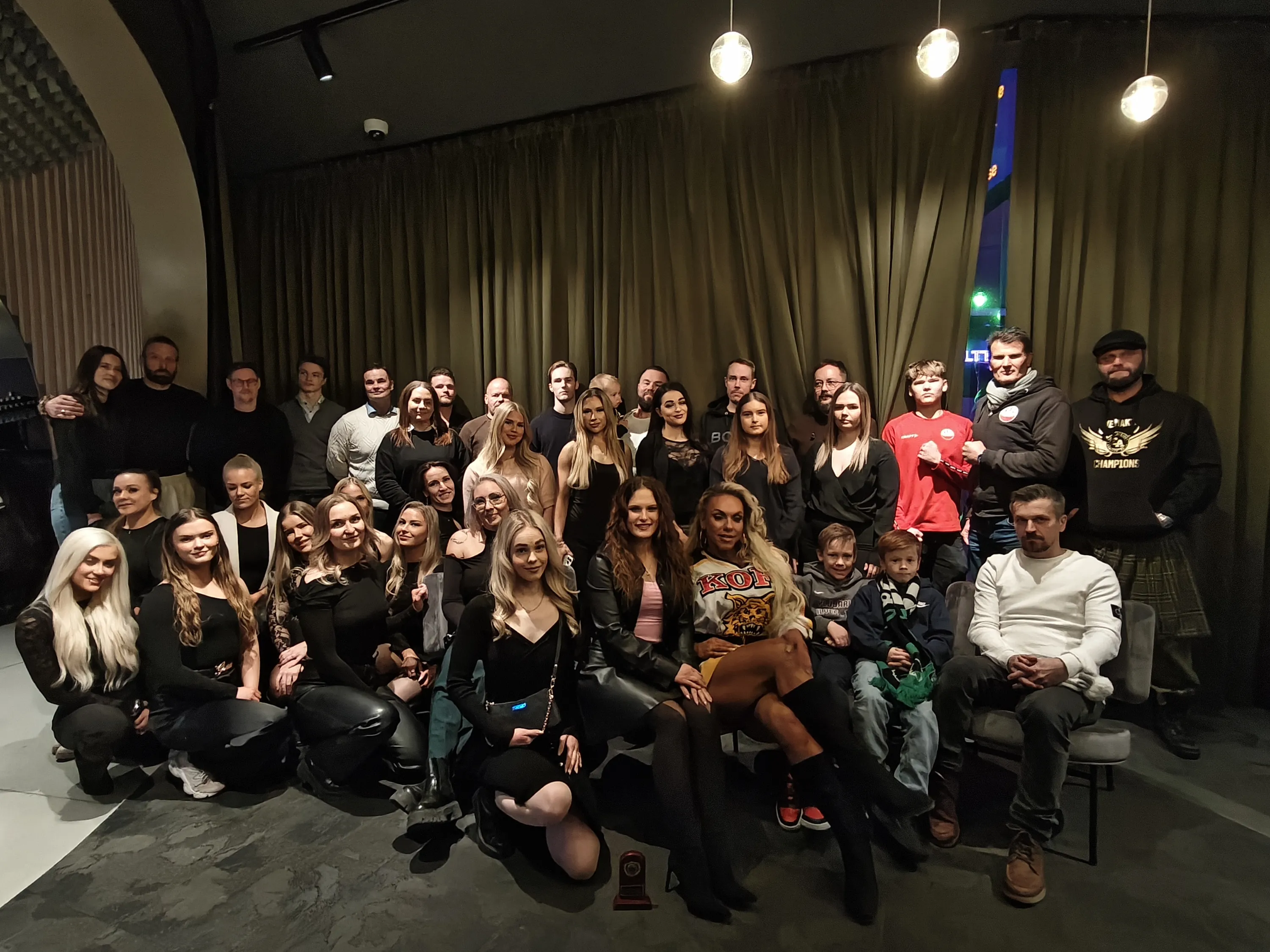 Suuri ryhmä ihmisiä, miehiä, naisia ja lapsia, poseeraa valokuvassa sisätiloissa. Taustalla on tummat verhot ja vasemmalla puolella puurimaseinä sekä akustiikkapaneeli. Katossa on kohdevaloja, riippuvalaisimia ja valvontakamera. Vasemmalla näkyy sininen pystysuora valaisin oransseilla numeroilla. Ihmiset ovat pukeutuneet arkisiin ja puolimuodollisiin vaatteisiin, enimmäkseen tummiin sävyihin. Osa seisoo, osa istuu tuoleilla ja osa polvistuu eturivissä. He katsovat kameraan ja hymyilevät. Lattialla oikealla on pieni palkinto. Valaistus on hämärä ja tunnelmallinen.