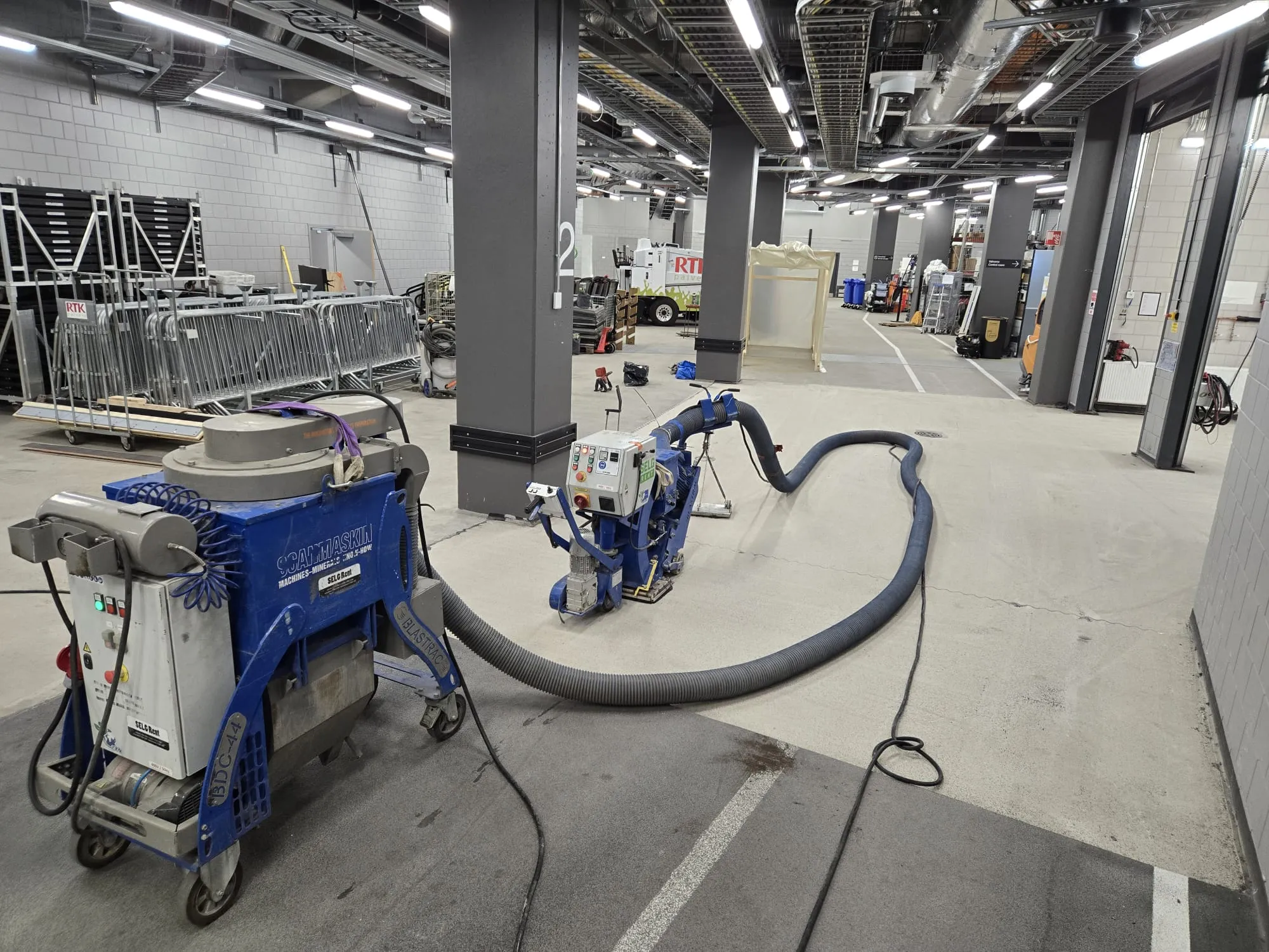 Kaksi sinistä teollisuuskonetta, todennäköisesti lattianpuhdistukseen tai hiekkapuhallukseen, on yhdistetty pitkällä harmaalla letkulla suuressa, valoisassa teollisuushallissa. Lattiat ovat betonia, ja seinät ovat harmaata tiiltä. Katossa on näkyvillä putkistoja ja kirkkaita loisteputkivaloja. Vasemmalla on pinottuna metalliaitoja ja mustia esteitä, ja taustalla näkyy valkoinen kuorma-auto. Lattialla on likaa ja musta sähkökaapeli.
