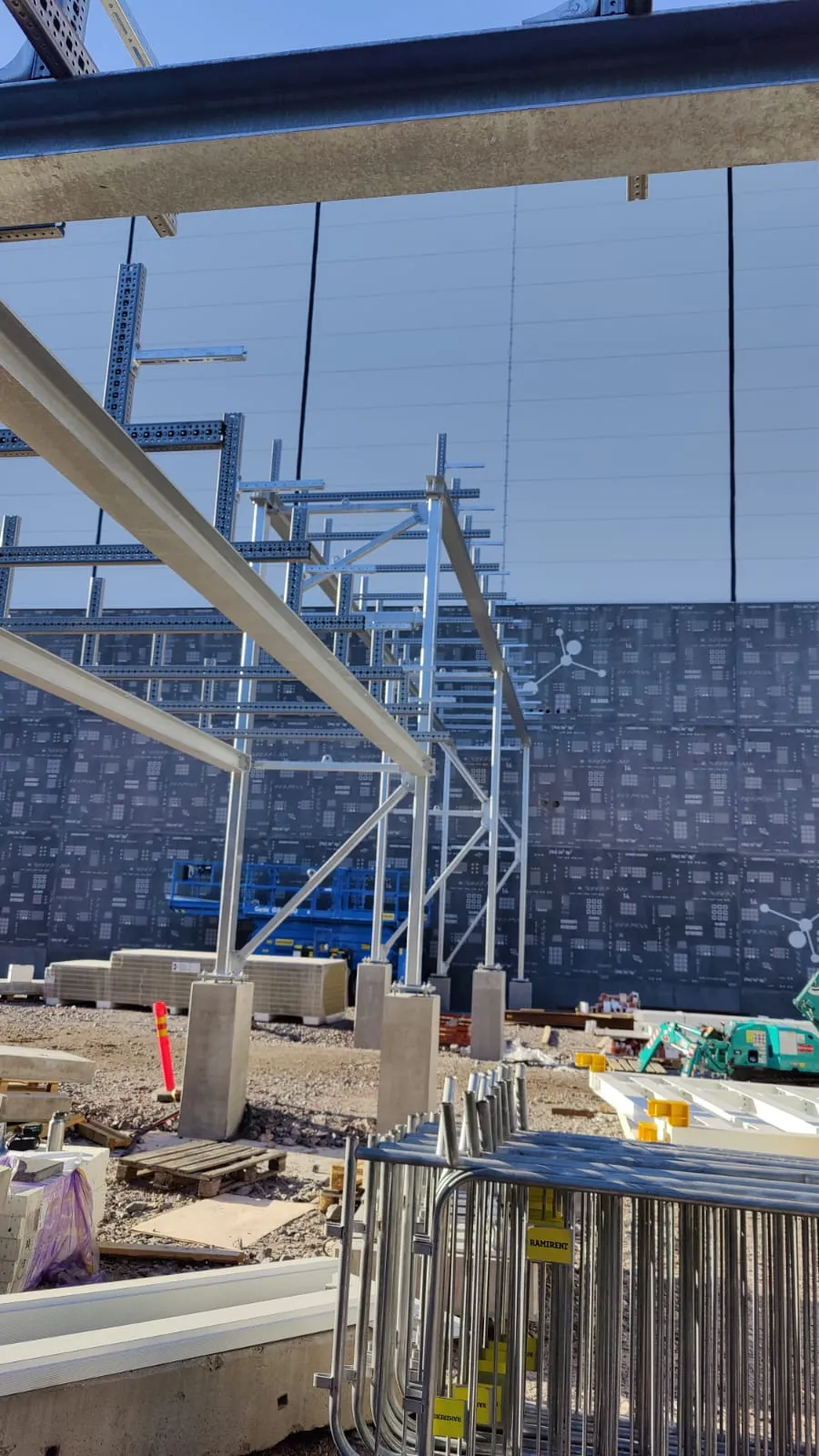 Aurinkoinen kuva rakennustyömaasta. Etualalla on pinottuja metalliaitoja ja soraa. Keskellä on monimutkainen hopeanvärinen metallirunko, joka koostuu palkeista ja tukirakenteista, tuettuna betonipilareilla. Taustalla näkyy tummanharmaa rakennuksen seinä, jossa on toistuva vaalea kuviointi, sekä vaaleampi seinä ja kirkas sininen taivas. Sininen saksinostin on näkyvissä metallirungon takana.