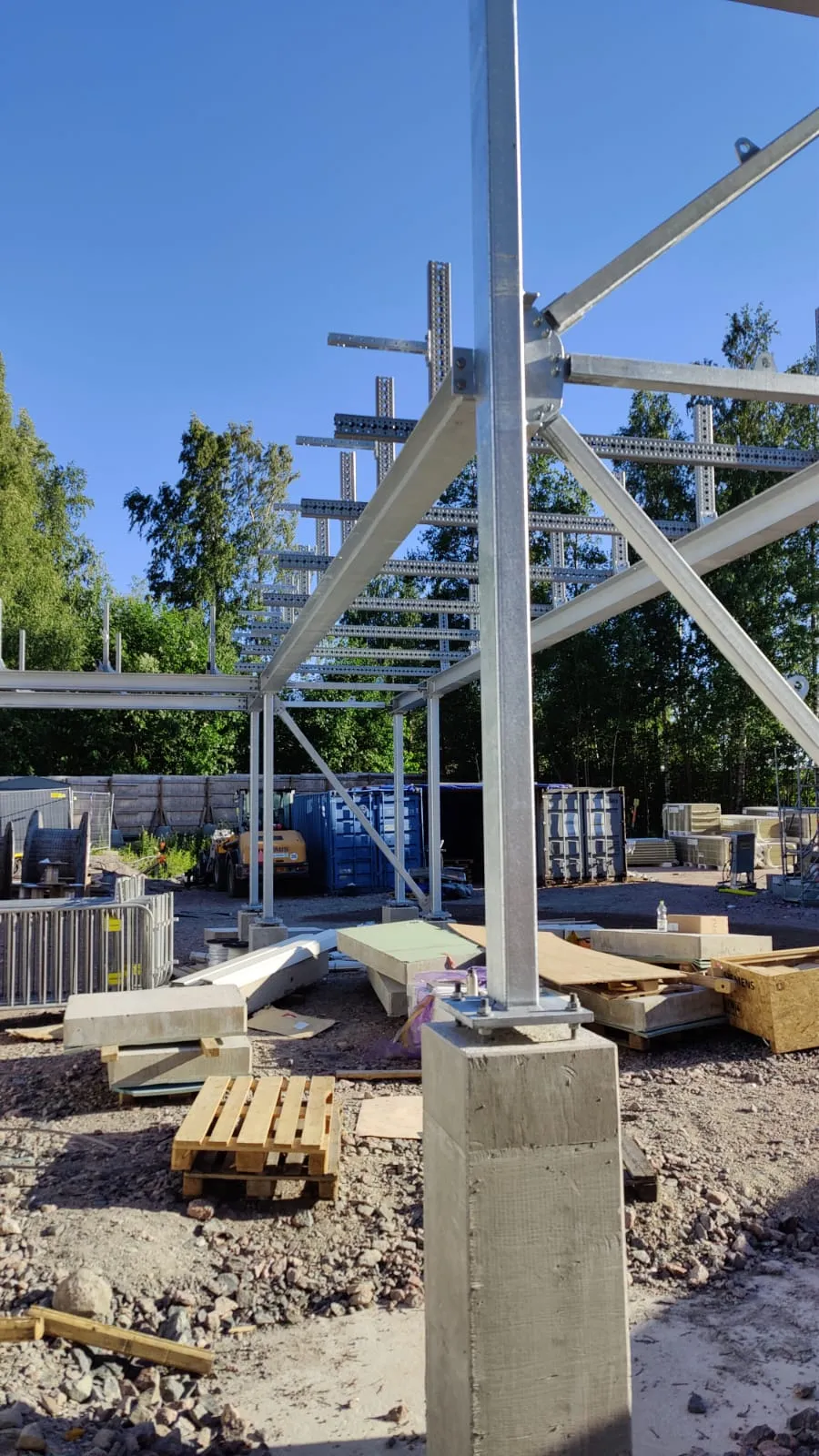 Aurinkoinen kuva rakennustyömaasta, jossa on osittain pystytetty galvanoitu teräsrunkorakenne. Etualalla on pystysuora teräspilari kiinnitettynä betonijalustaan. Taustalla näkyy sininen taivas, vihreitä puita, sinisiä merikontteja, keltainen työkone ja erilaisia rakennustarvikkeita kivisellä maalla.