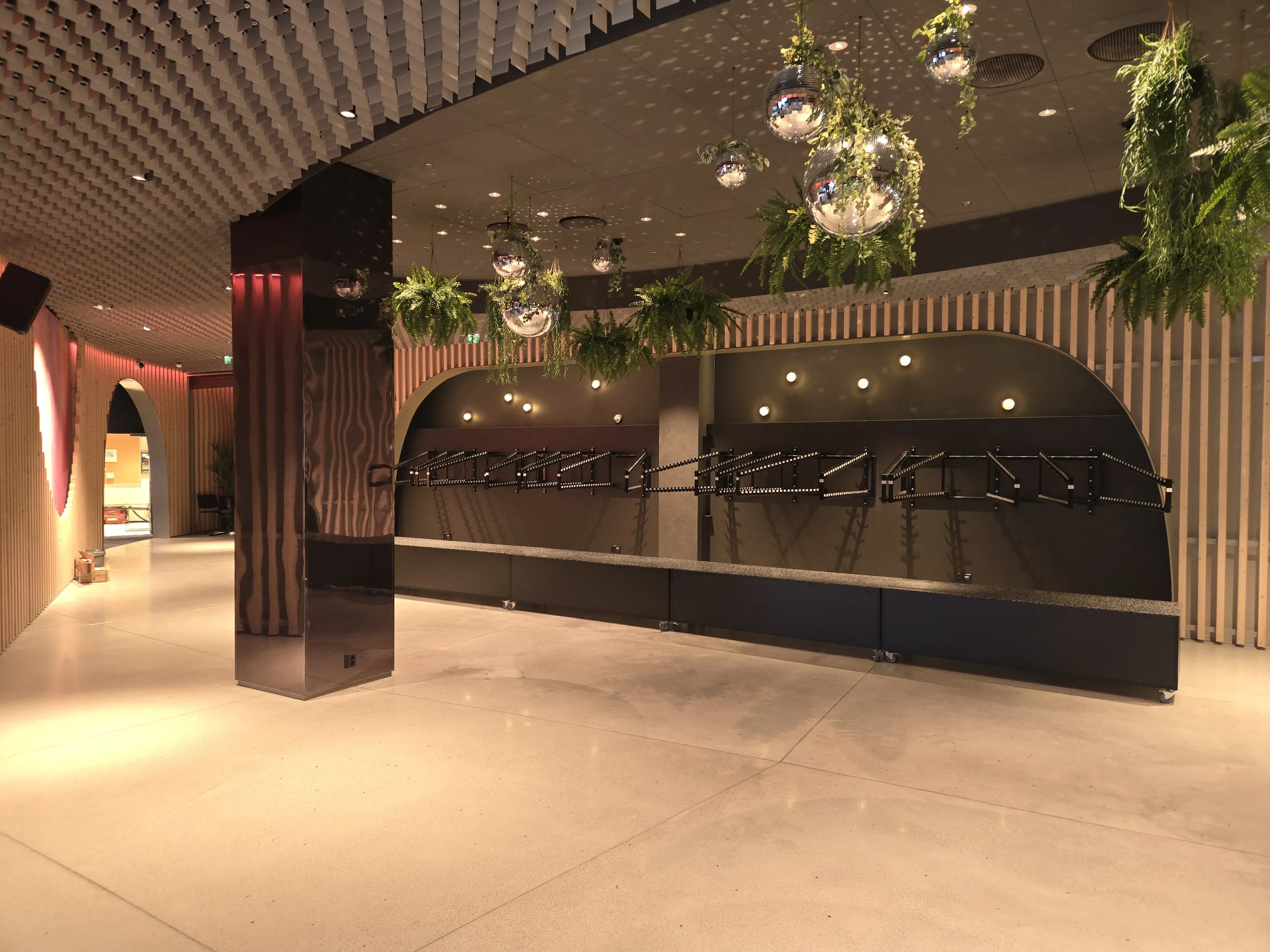 Sisäkuva modernista tilasta, jossa on vaalea betonilattia ja tumma katto. Vasemmalla on puurimaseinäinen käytävä, jossa on kaareva aukko ja heijastava tumma pilari. Käytävän katossa on kuvioitu paneelointi. Oikealla seinällä on tumma kaareva syvennys, jossa on useita mustia telineitä ja pyöreitä valaisimia. Yläpuolella roikkuu viherkasveilla ympäröityjä peilipalloja, jotka heijastavat valoa tummaan kattoon. Katossa on myös upotettuja valoja ja ilmanvaihtoritilöitä. Tilan valaistus on lämmin ja luo tunnelmallisen ilmapiirin.