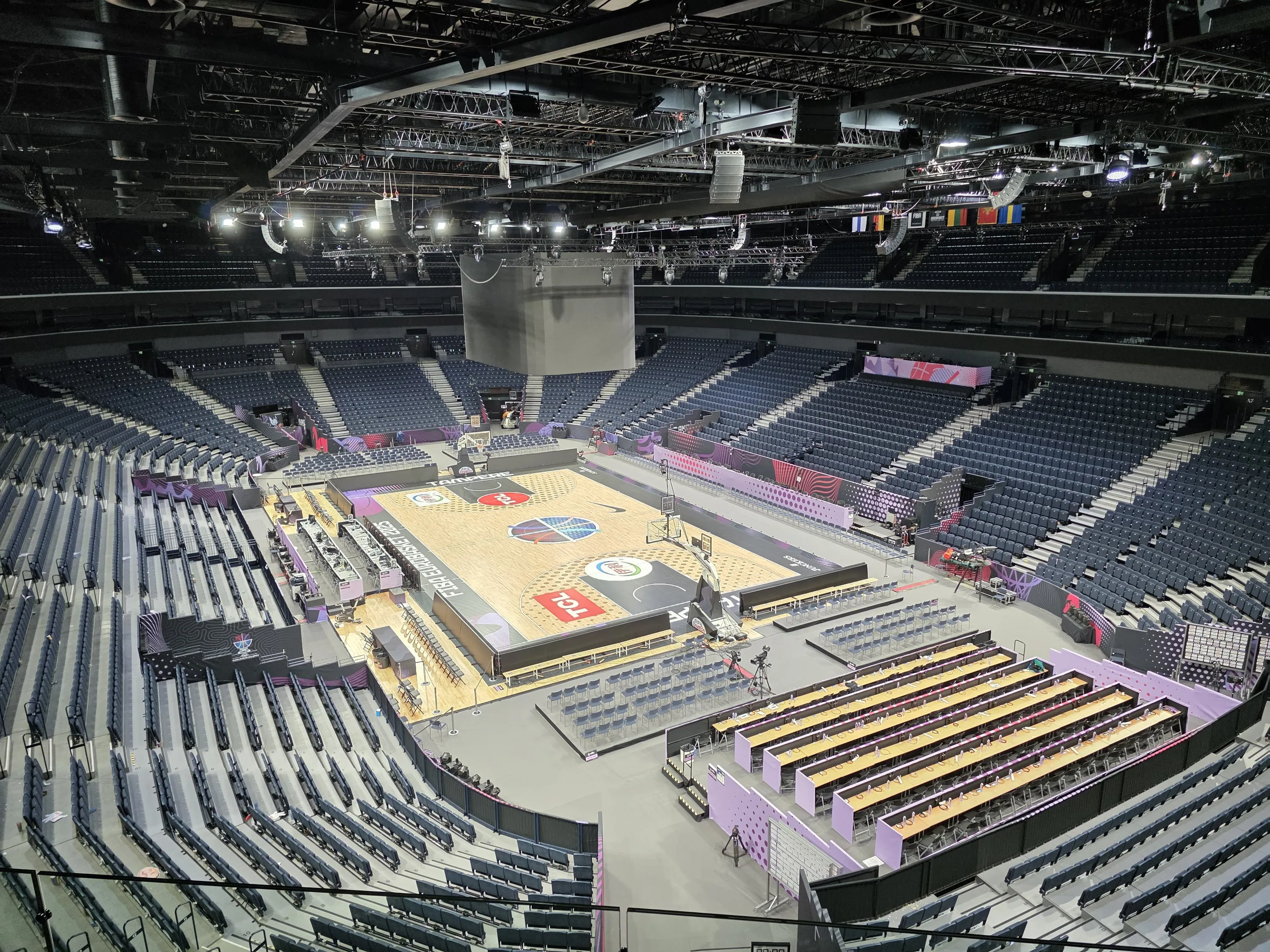 Ylhäältä otettu kuva tyhjästä sisäkoripalloareenasta, jossa on vaalea puinen kenttä FIBA EuroBasket 2025 -logoilla. Tummanharmaat istuimet ympäröivät kenttää useissa tasoissa. Keskellä katosta roikkuu suuri tumma näyttö, ja kattorakenteissa on valoja, kaiuttimia ja ilmanvaihtoputkia. Yhdellä sivulla on pitkiä pöytiä medialle ja useita kansallislippuja roikkuu katosta. Valaistus on kirkas kentän yllä, kun taas istuinalueet ovat hämärämpiä.